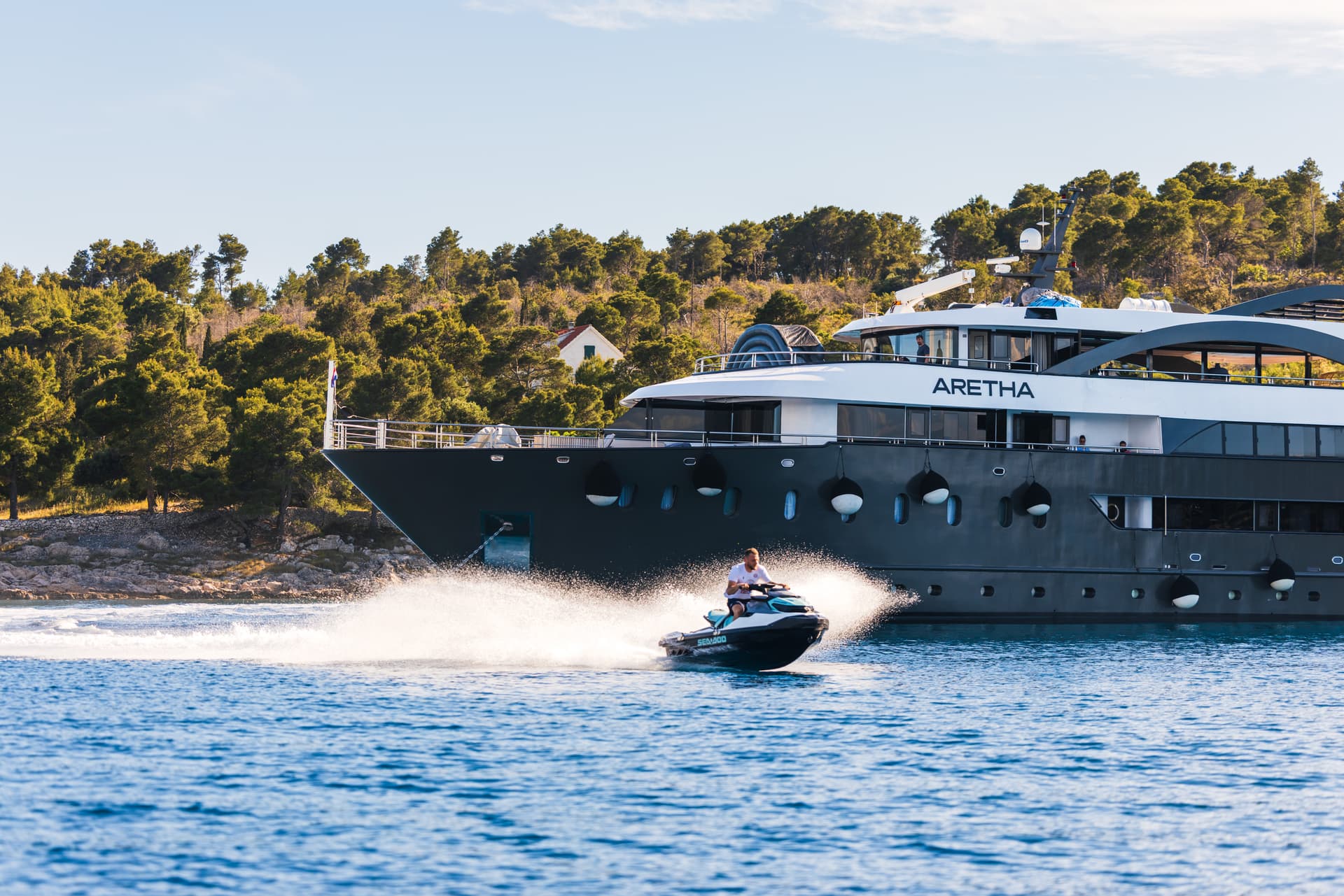Man on jet ski sprays water near large yacht named Aretha off wooded coastline.