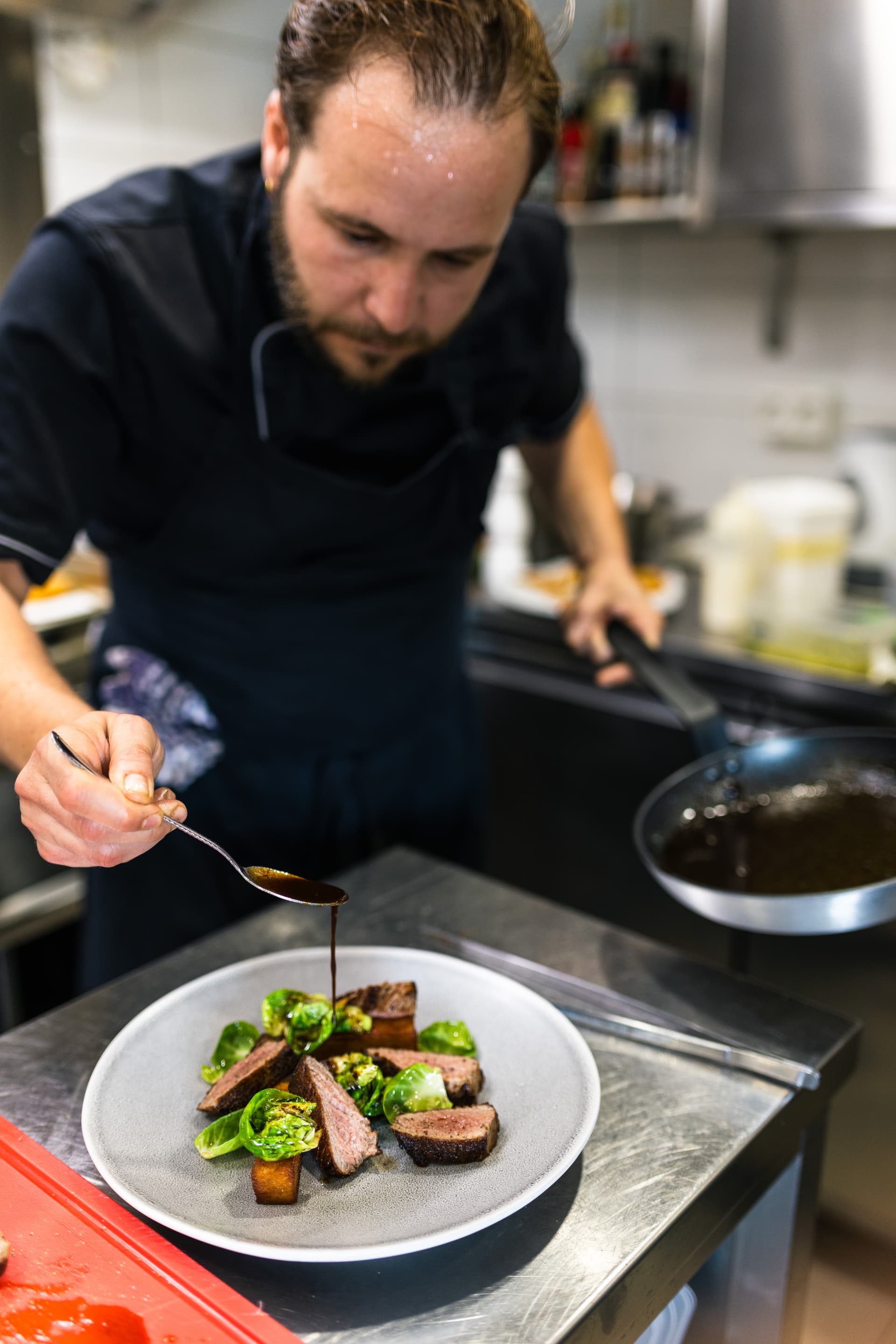 Chef plating sliced steak with sauce, Brussels sprouts, and potatoes in a commercial kitchen.