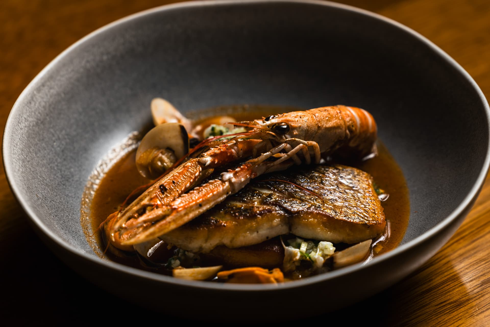 Seared fish fillet with langoustine and shellfish in brown broth served in a gray bowl.