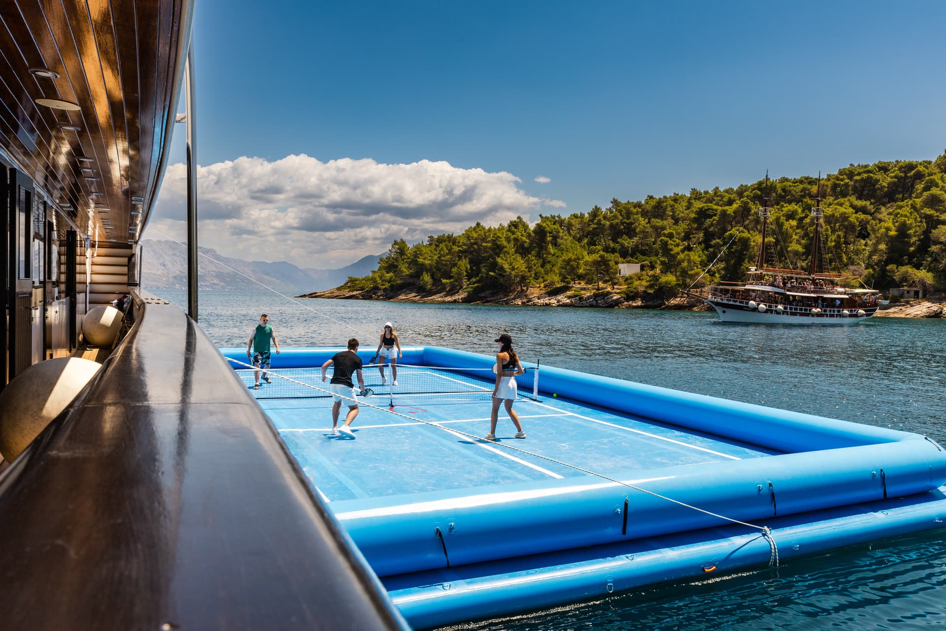 People playing pickleball on an inflatable court floating on blue water near a forested coastline.