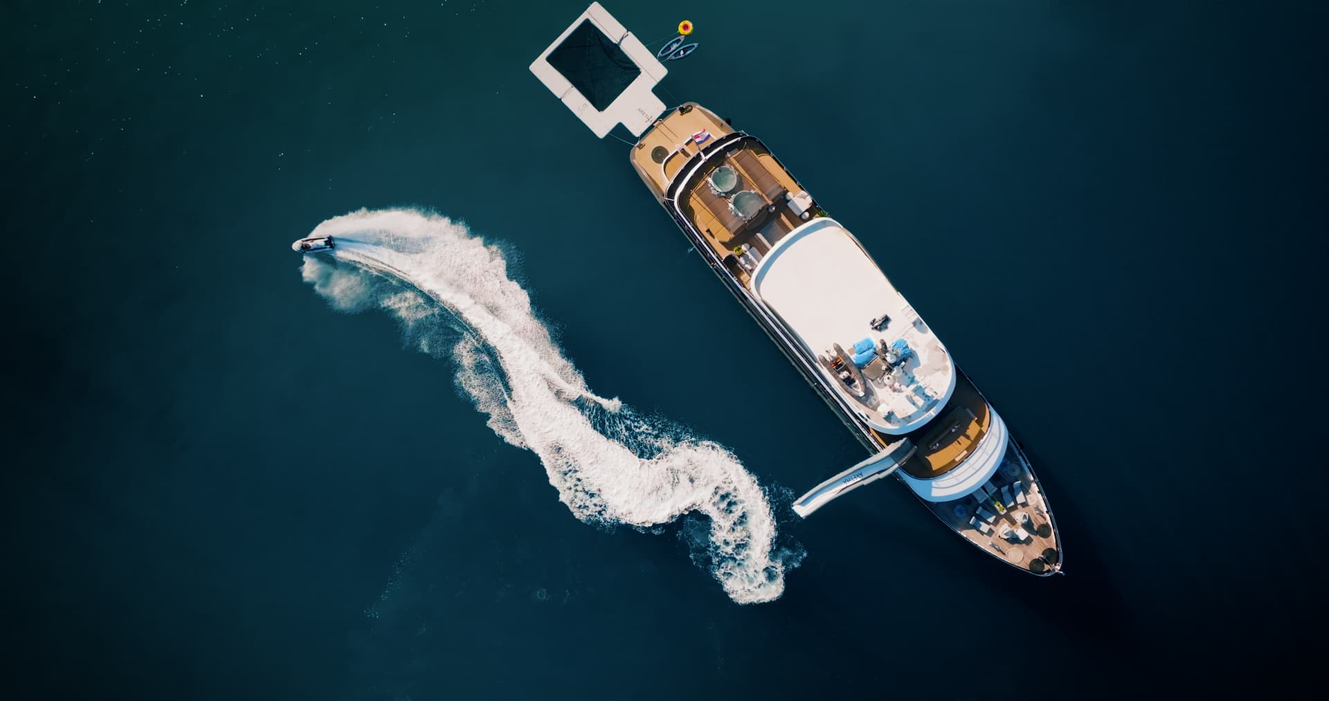 Aerial view of luxury yacht with water slide and jet ski creating white wake on dark blue sea.