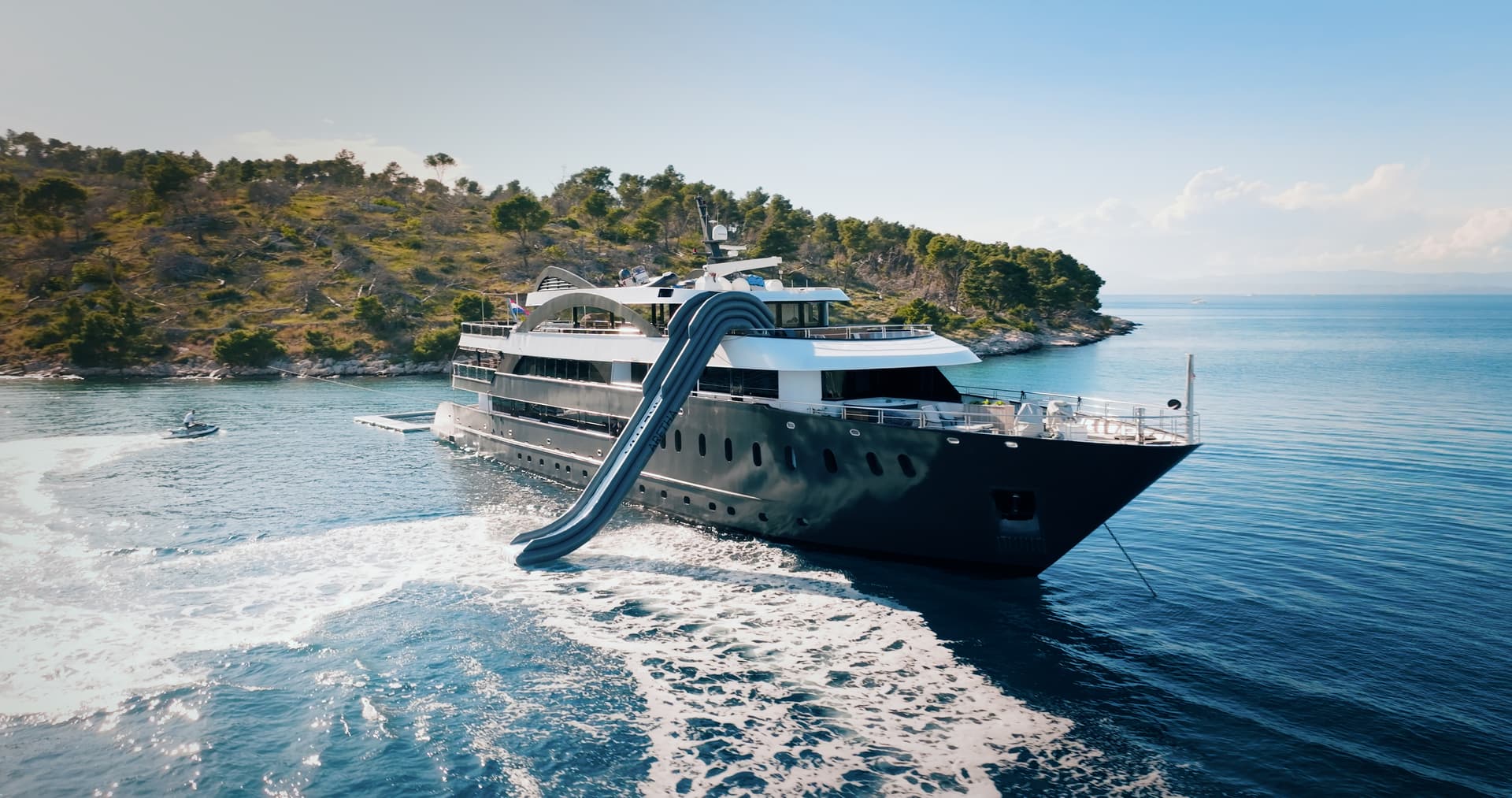 Large yacht with an inflatable water slide anchored near a wooded coastline on blue sea.