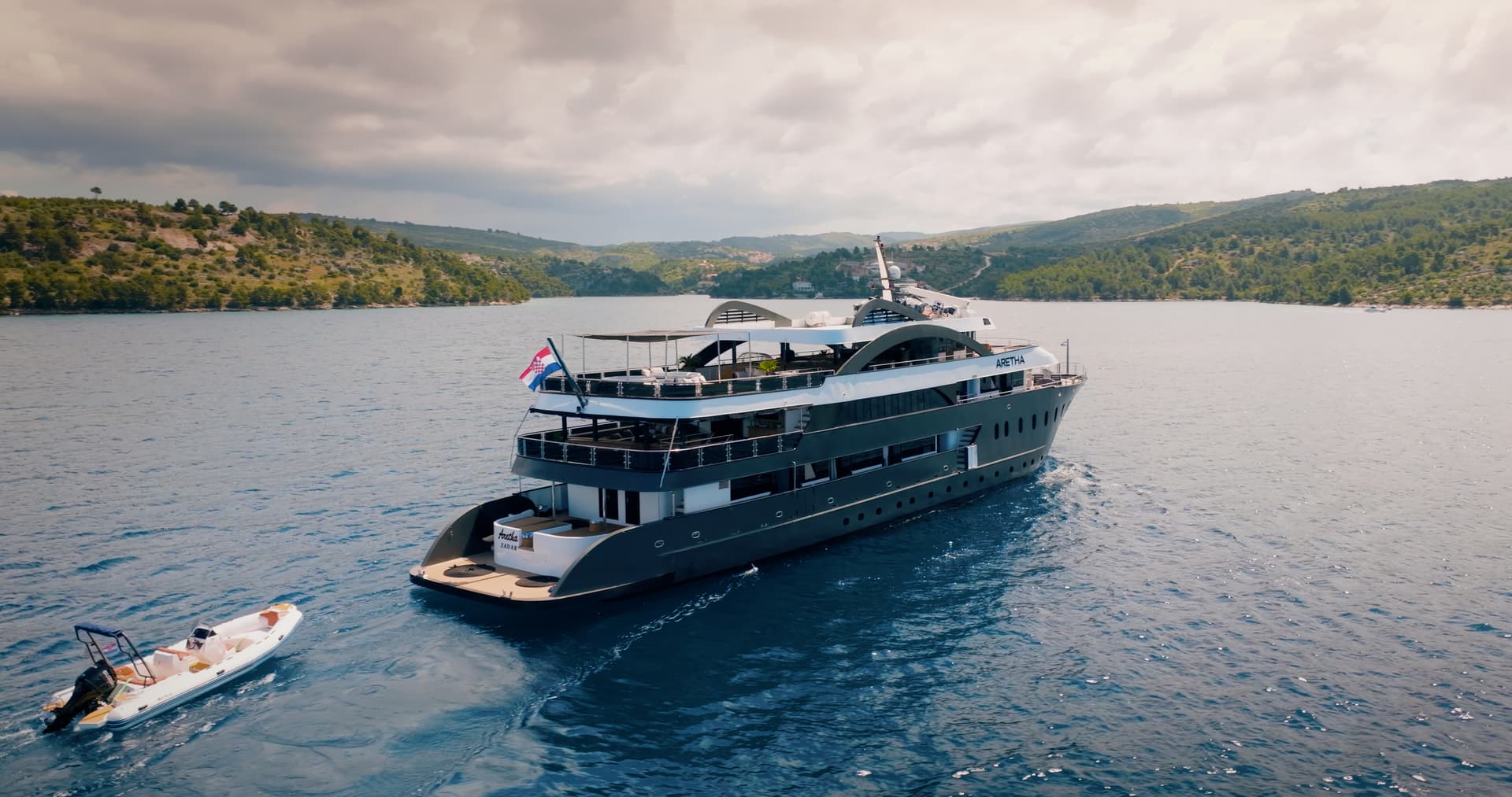 Large yacht named Aretha with Croatian flag towing a small inflatable boat near forested coastline.