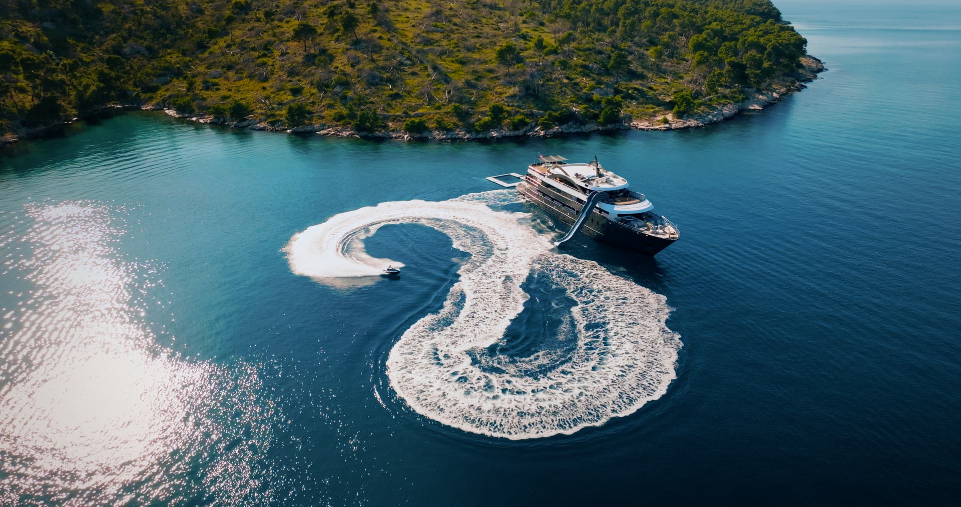 Yacht anchored near forested coastline while jet ski creates white wake on deep blue water.