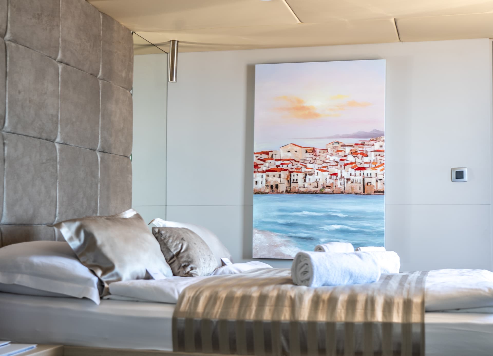 Hotel room with neatly made bed, plush pillows, and painting of coastal town at sunset.