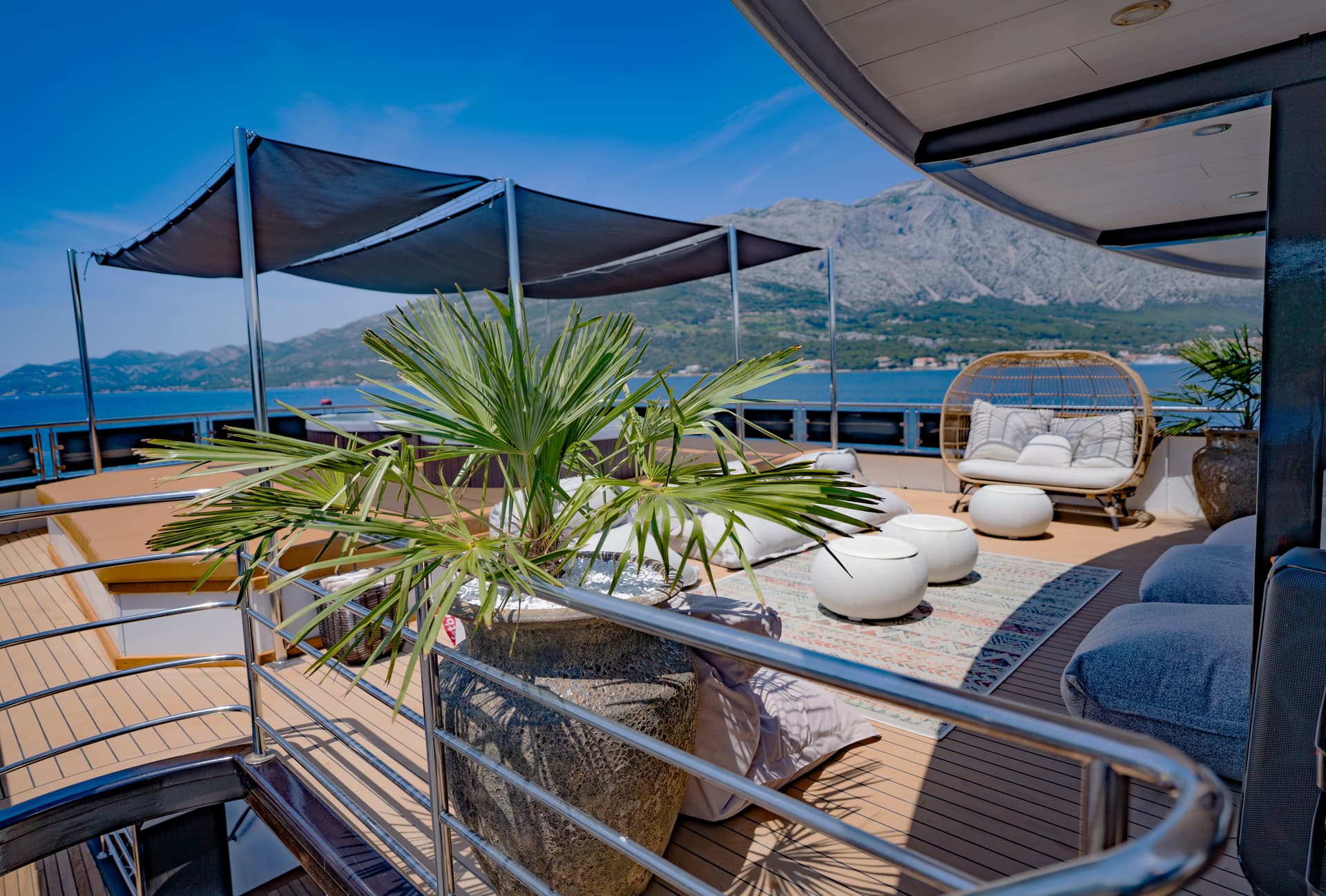 Yacht deck lounge with palm plant, wicker chair, and mountains across blue sea.