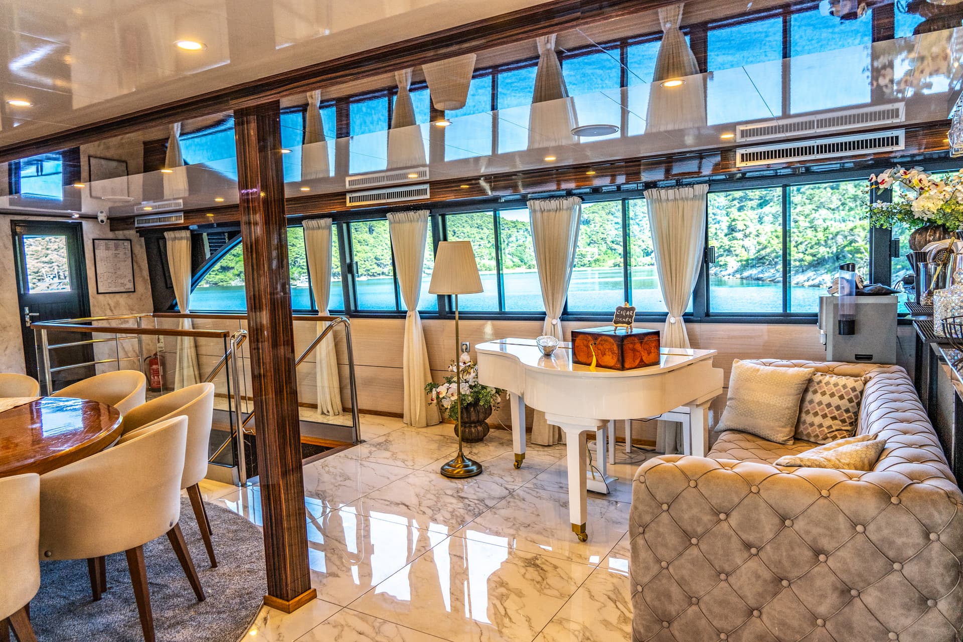 Luxury yacht lounge with white piano, tufted sofa, and views of forested coastline.