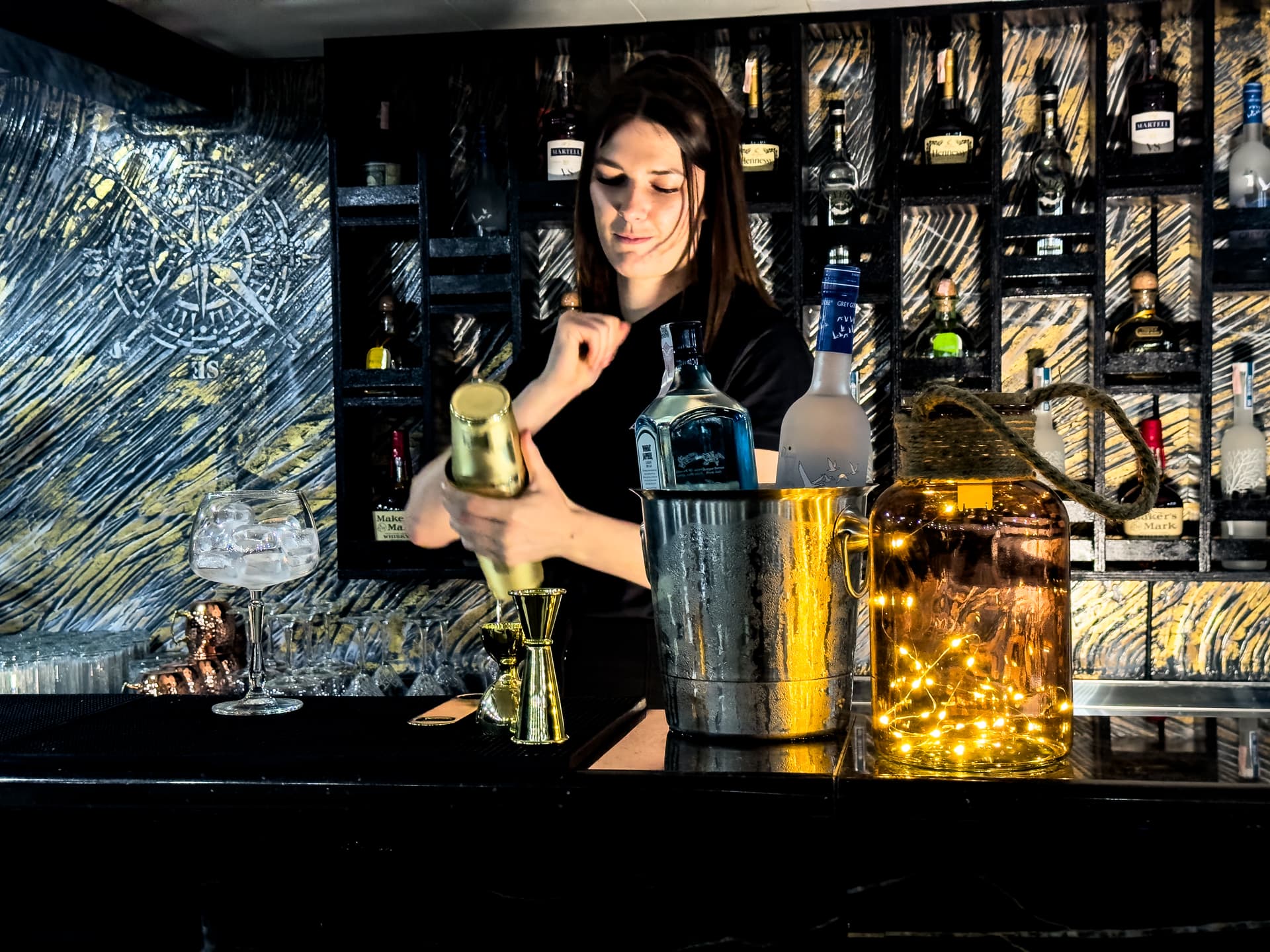 Bartender shaking cocktail in gold shaker at dimly lit bar with liquor bottles displayed.