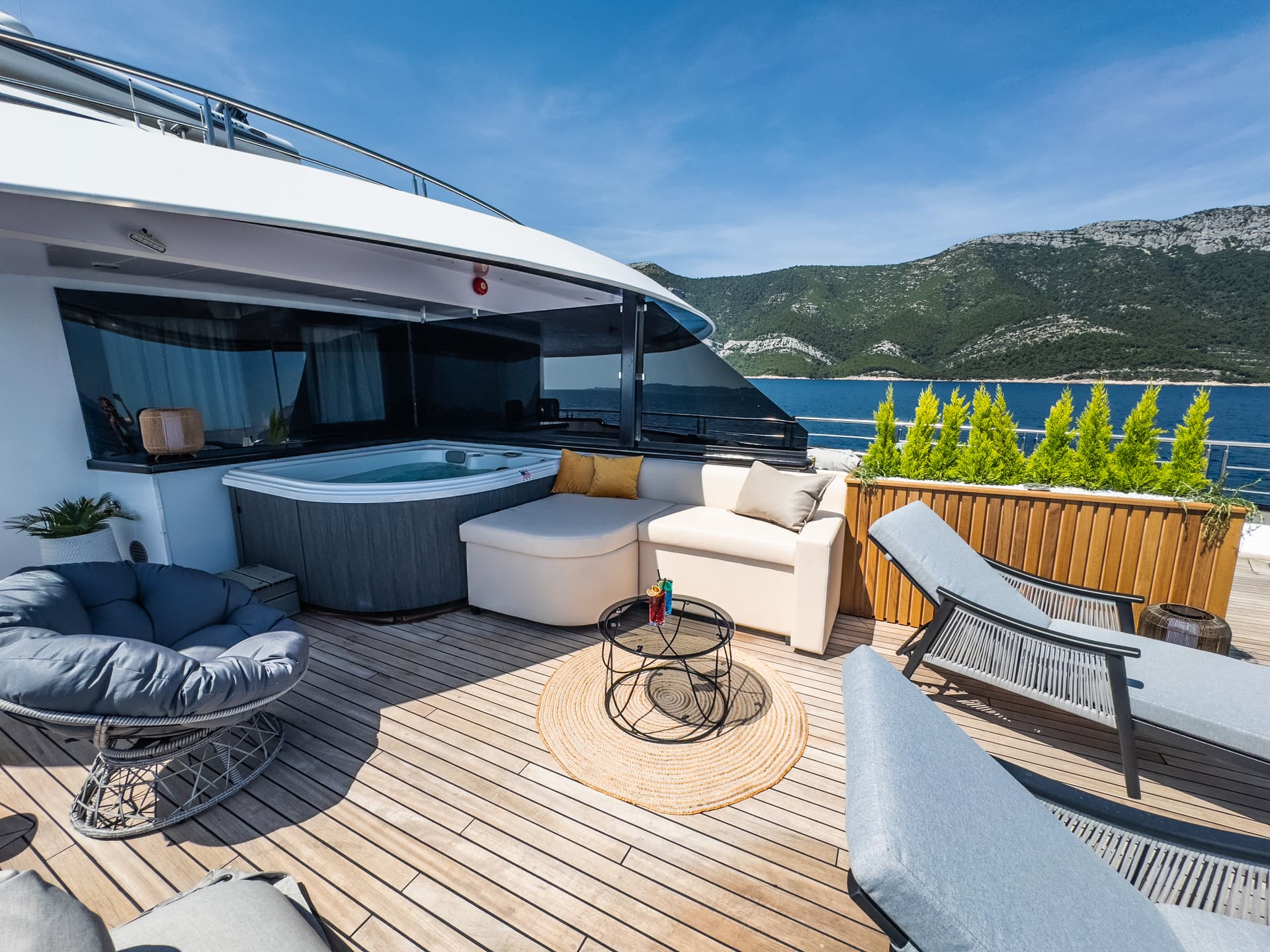 Luxury yacht deck with hot tub, lounge seating, and view of green, rocky coastline.