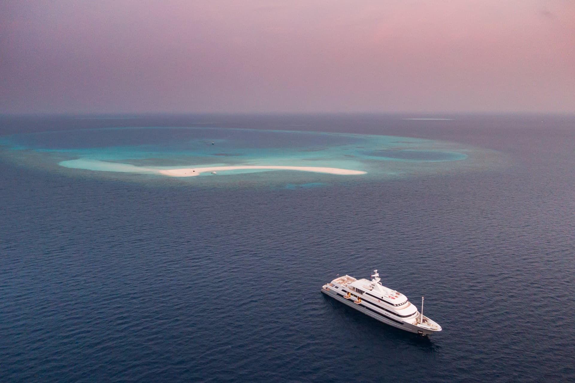 Large yacht floating on dark blue ocean near a shallow sandbar with turquoise water.