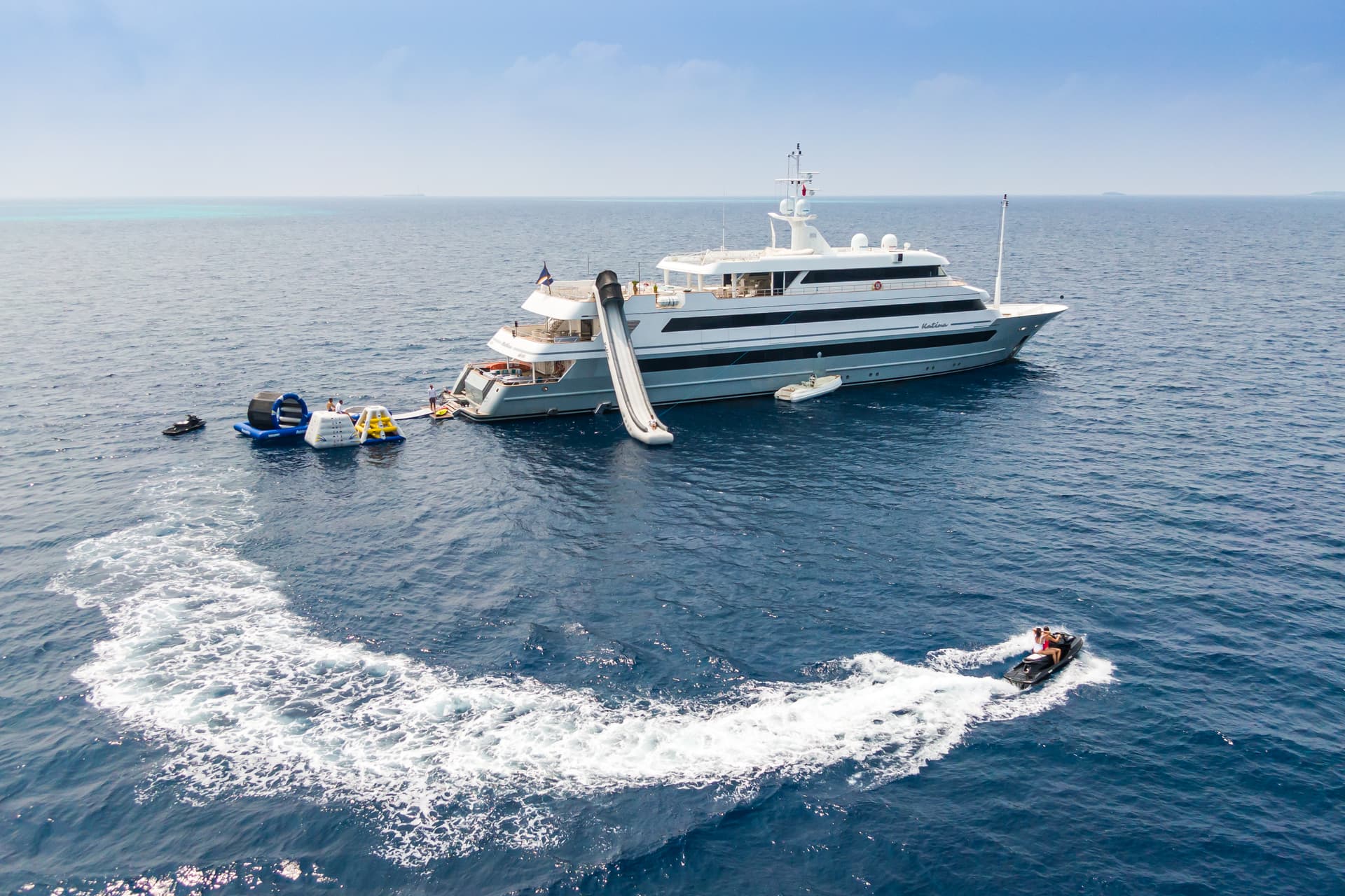 Luxury yacht anchored with water slide, inflatable toys, and jet ski activity on blue sea.