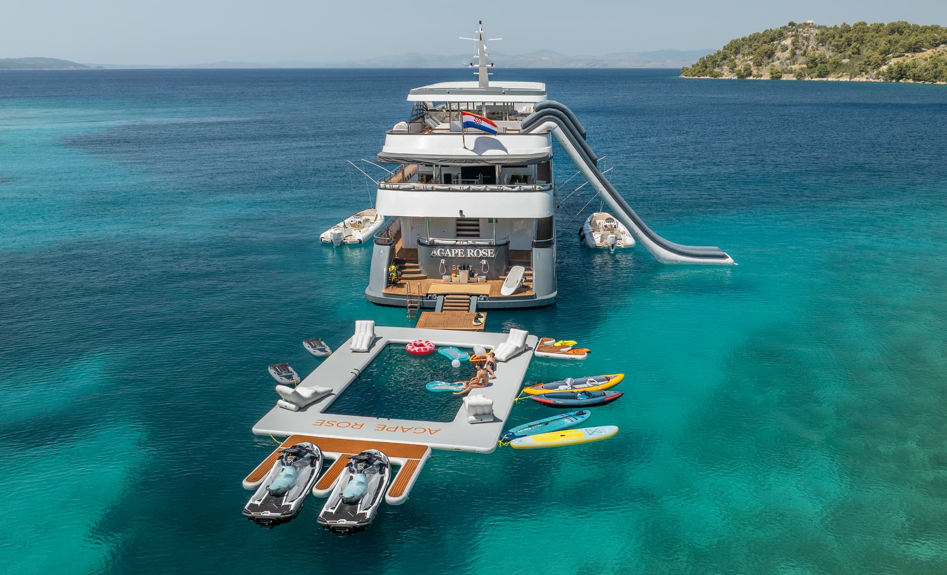 Large yacht anchored near a wooded island with an inflatable water park and jet skis in turquoise sea.