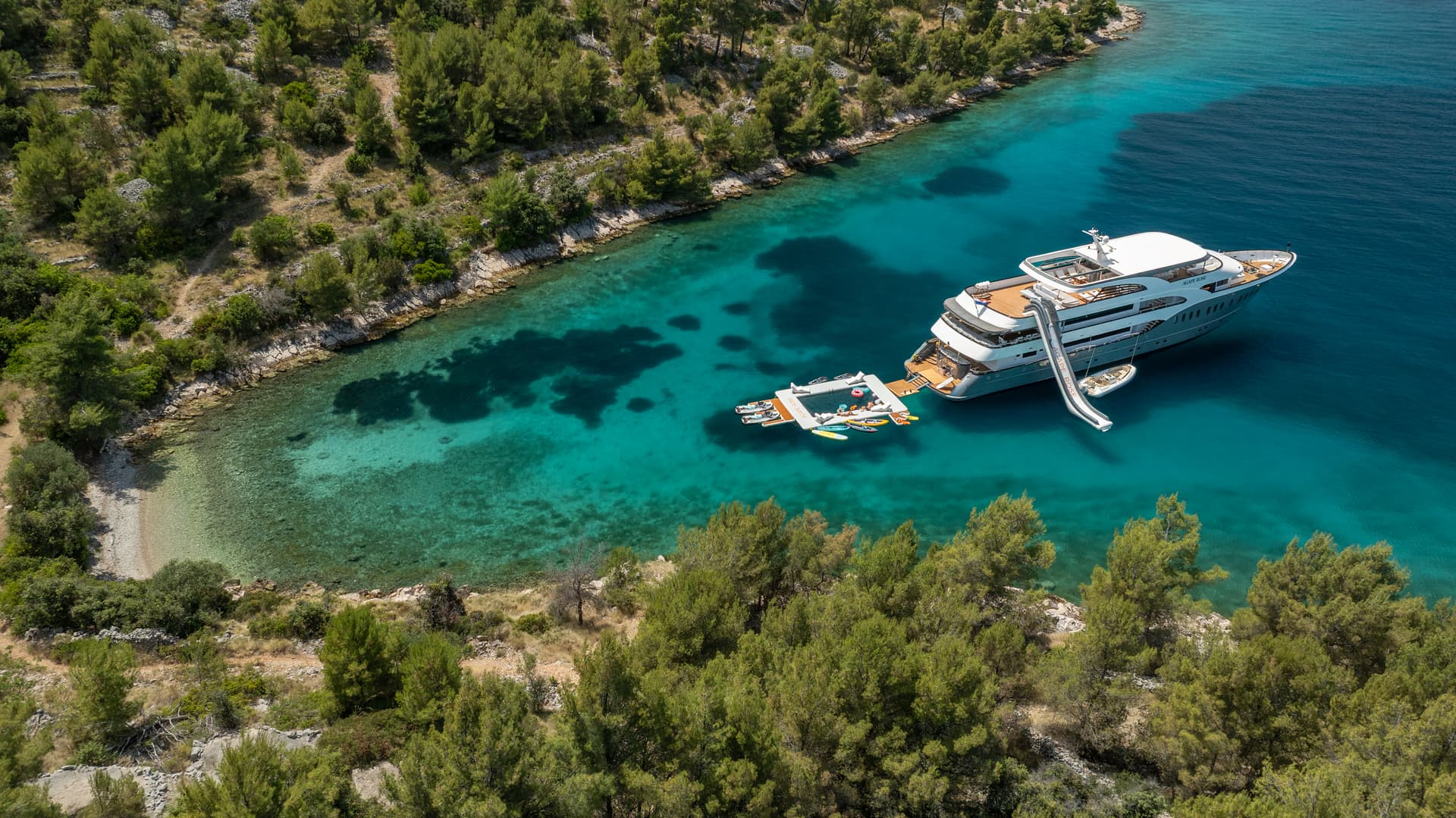 Luxury yacht anchored in cove with turquoise water, inflatable water toys, and slide.