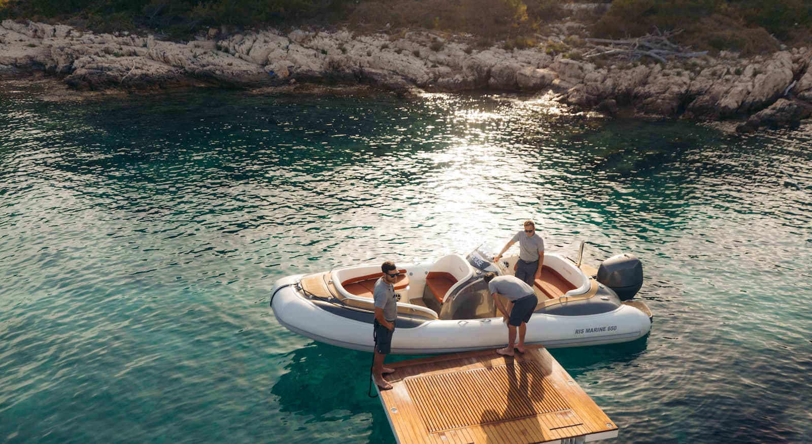Three people on a rigid inflatable boat next to a wooden dock near a rocky coastline.