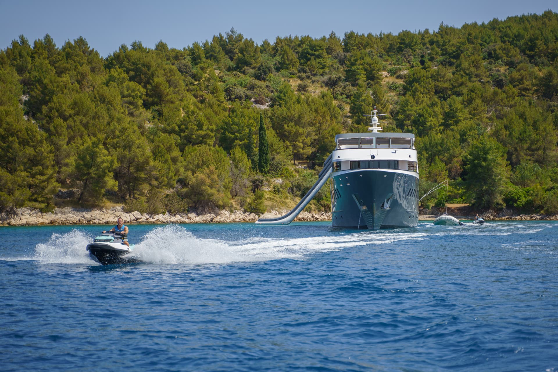 Man riding jet ski near large yacht anchored off wooded coastline.
