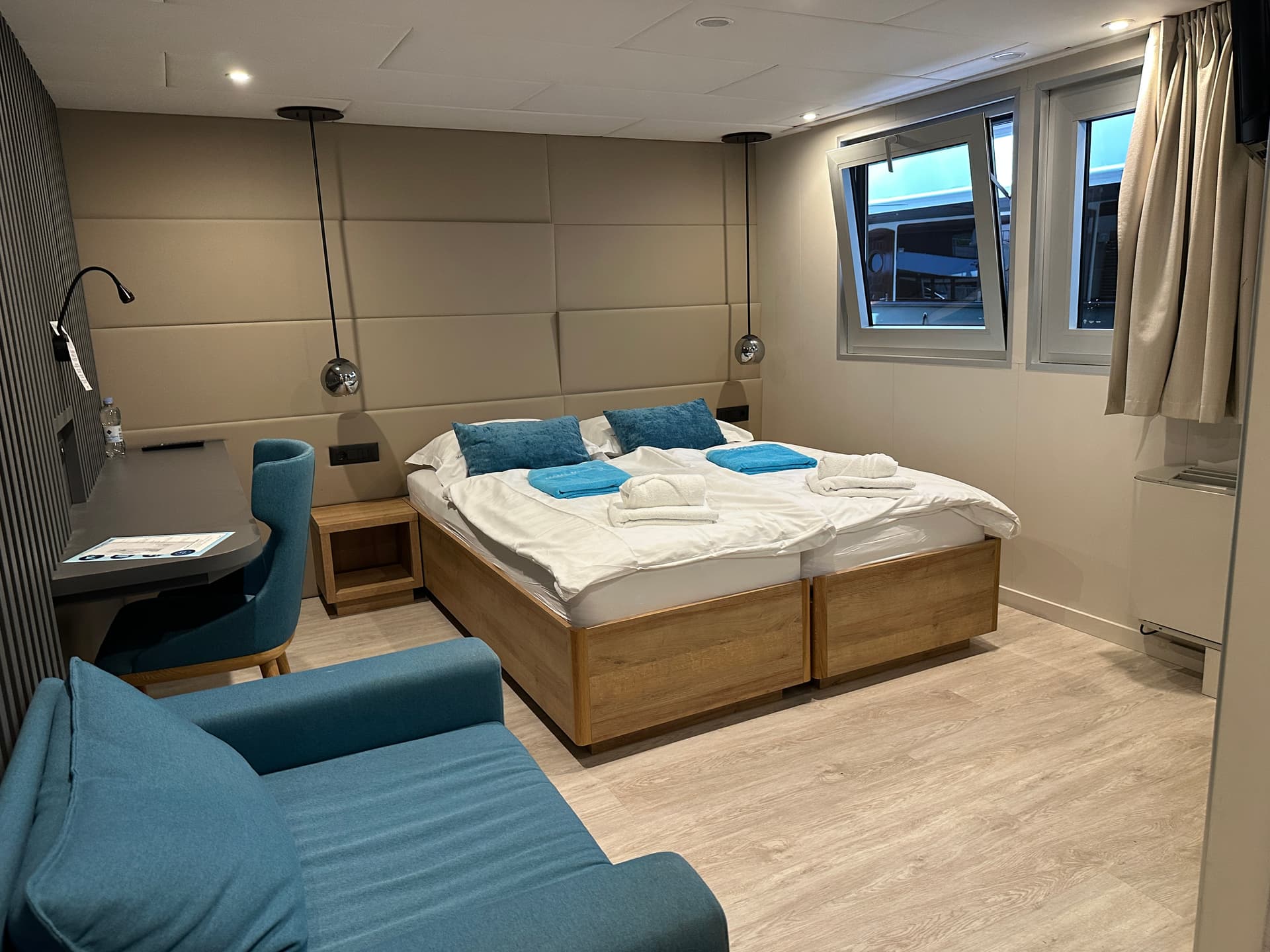 Cruise ship cabin interior with twin beds, blue cushions, desk, and armchair