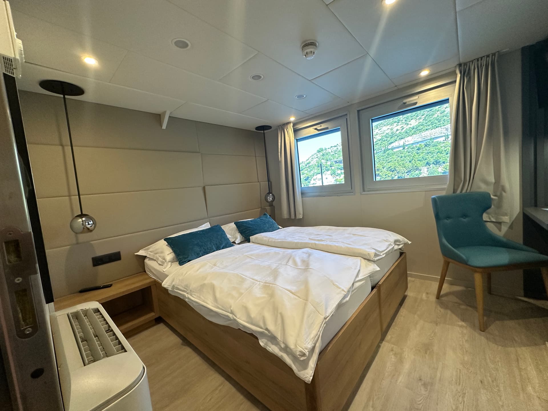 Cruise ship main deck cabin with twin beds, blue pillows, and windows showing outdoor scenery