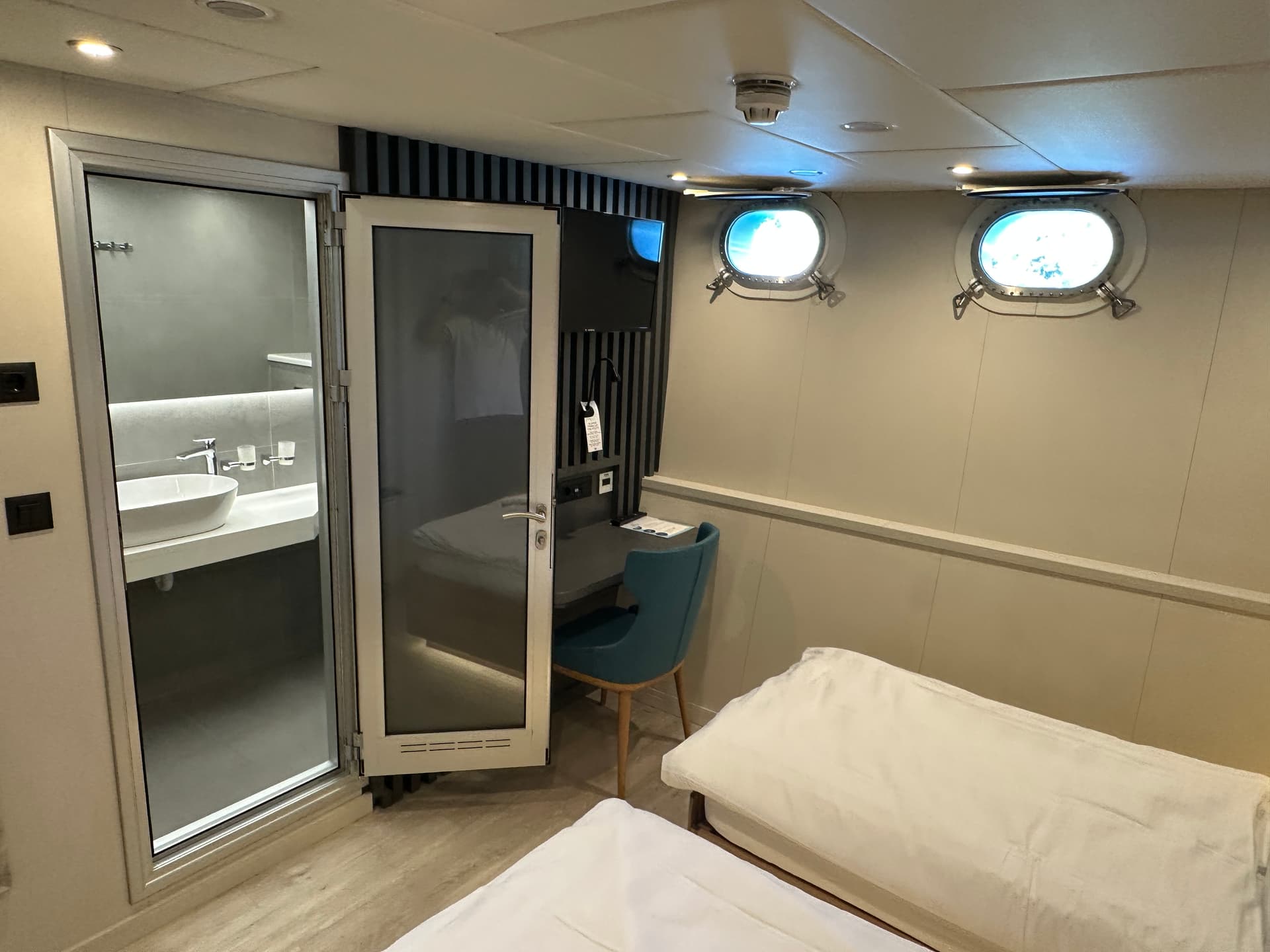 Standard ship cabin with twin beds, desk, and attached bathroom with portholes