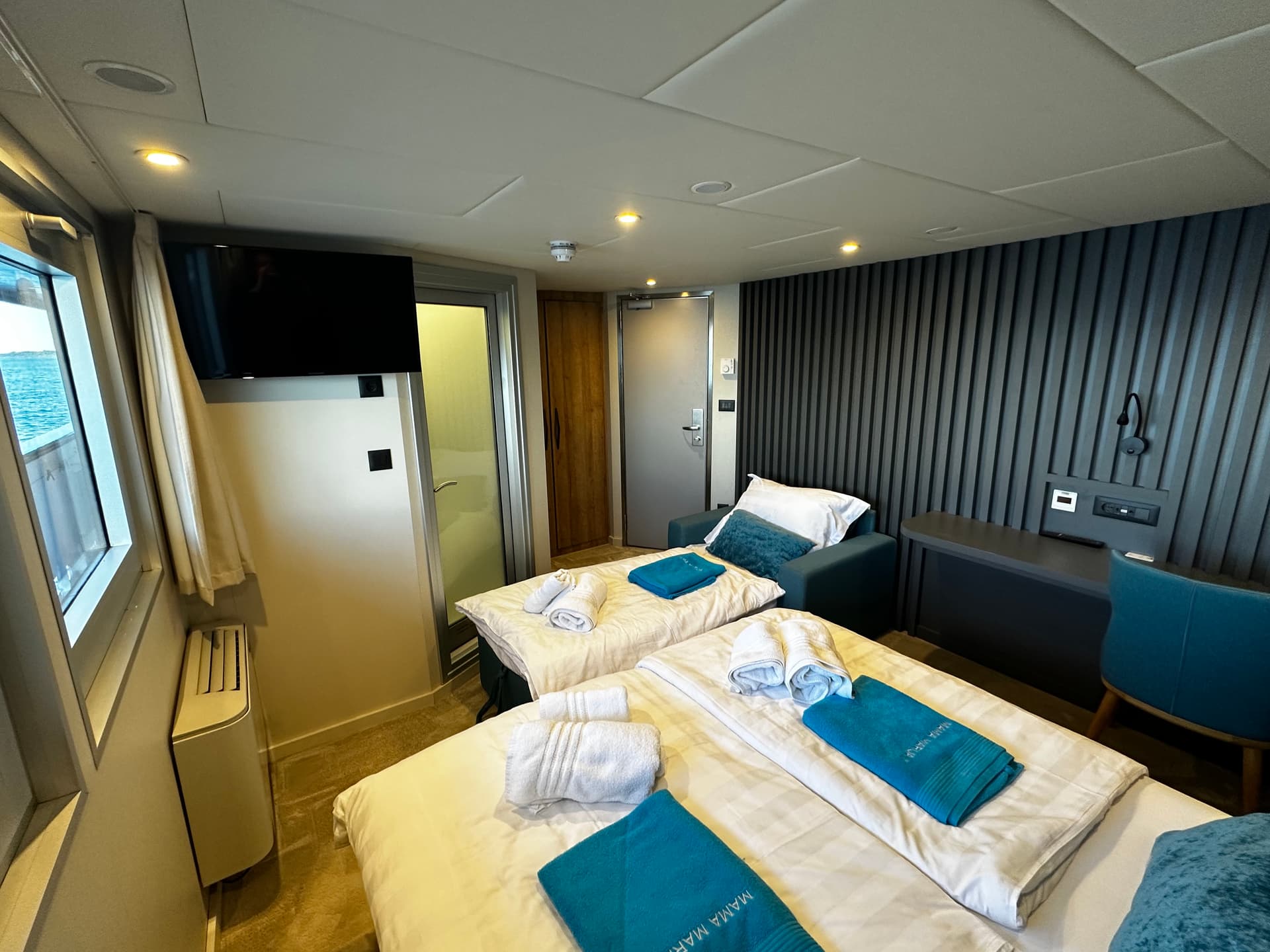 Modern cruise ship cabin interior with twin beds, folded towels, and ocean view window