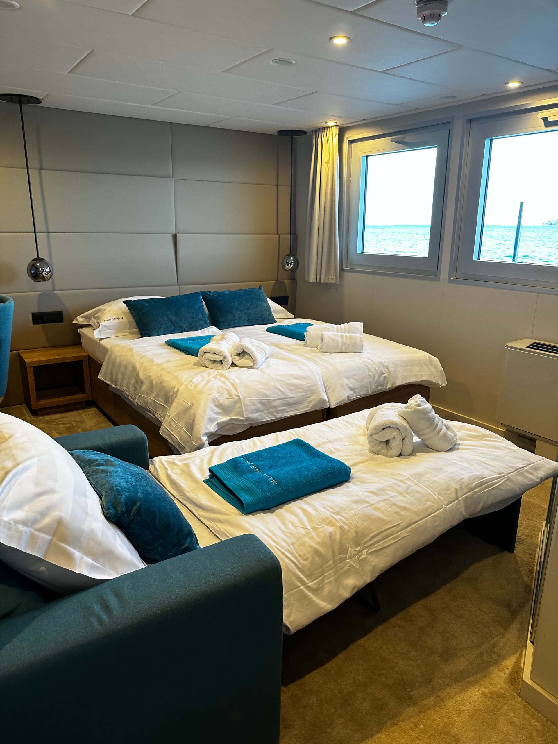 Modern cruise ship cabin with twin beds, blue pillows, rolled towels, and ocean-view windows
