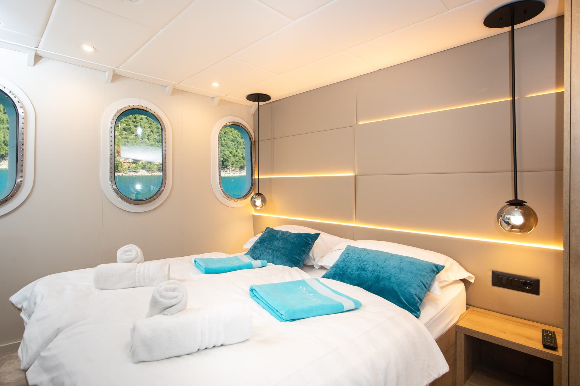 Modern ship cabin bedroom with white linens, turquoise pillows, rolled towels, and round porthole windows showing water an...