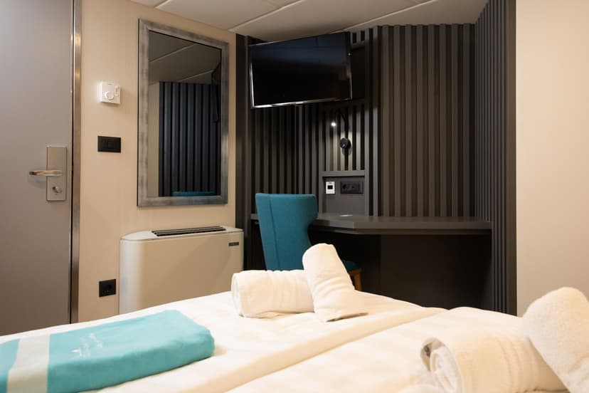 Ship cabin interior with bed, rolled white towels, turquoise chair, desk, wall-mounted television, and mirror