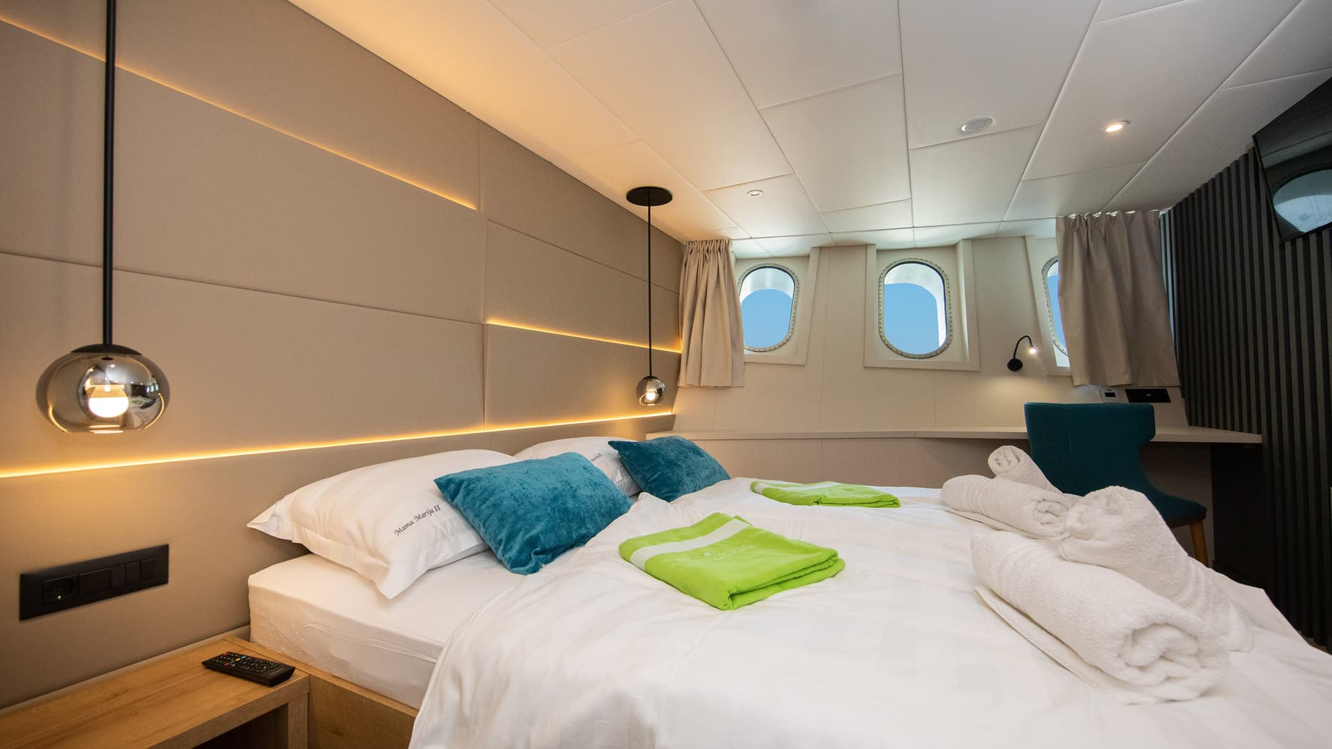Cabin bedroom with double bed, blue pillows, green towels, and portholes