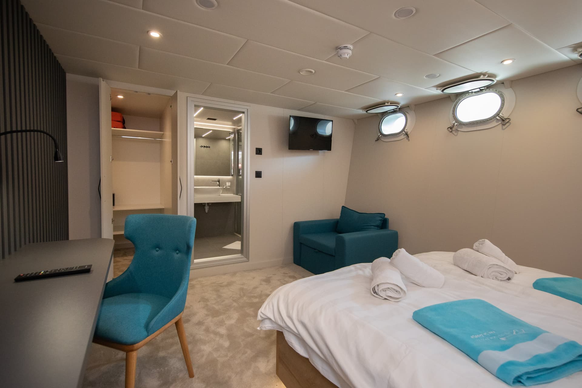 Modern standard cabin interior with bed, desk, blue chair, and ensuite bathroom