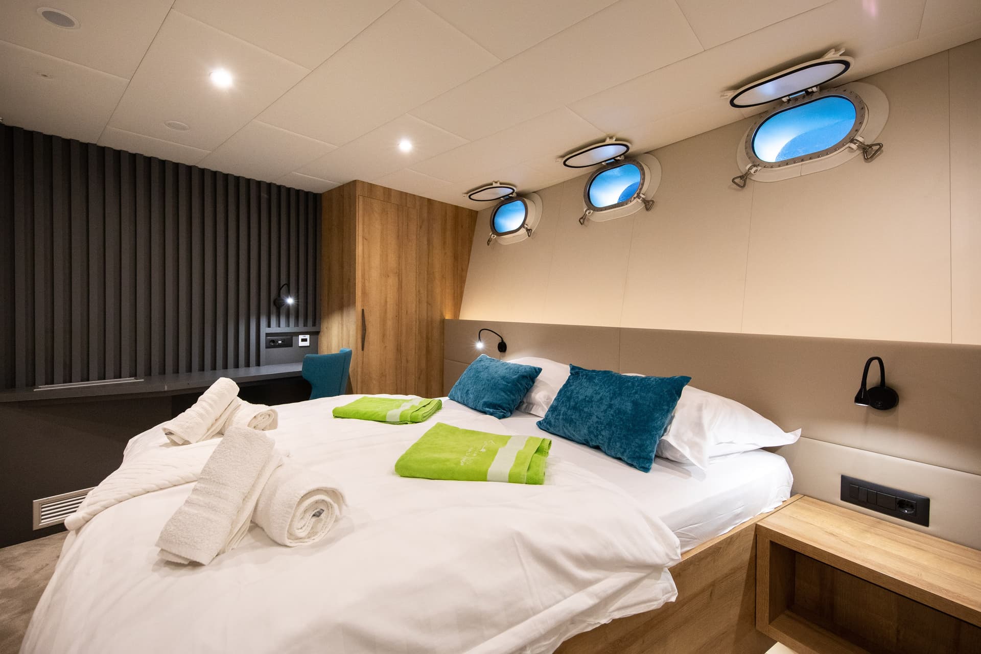 Modern yacht cabin with a double bed, rolled towels, and round portholes