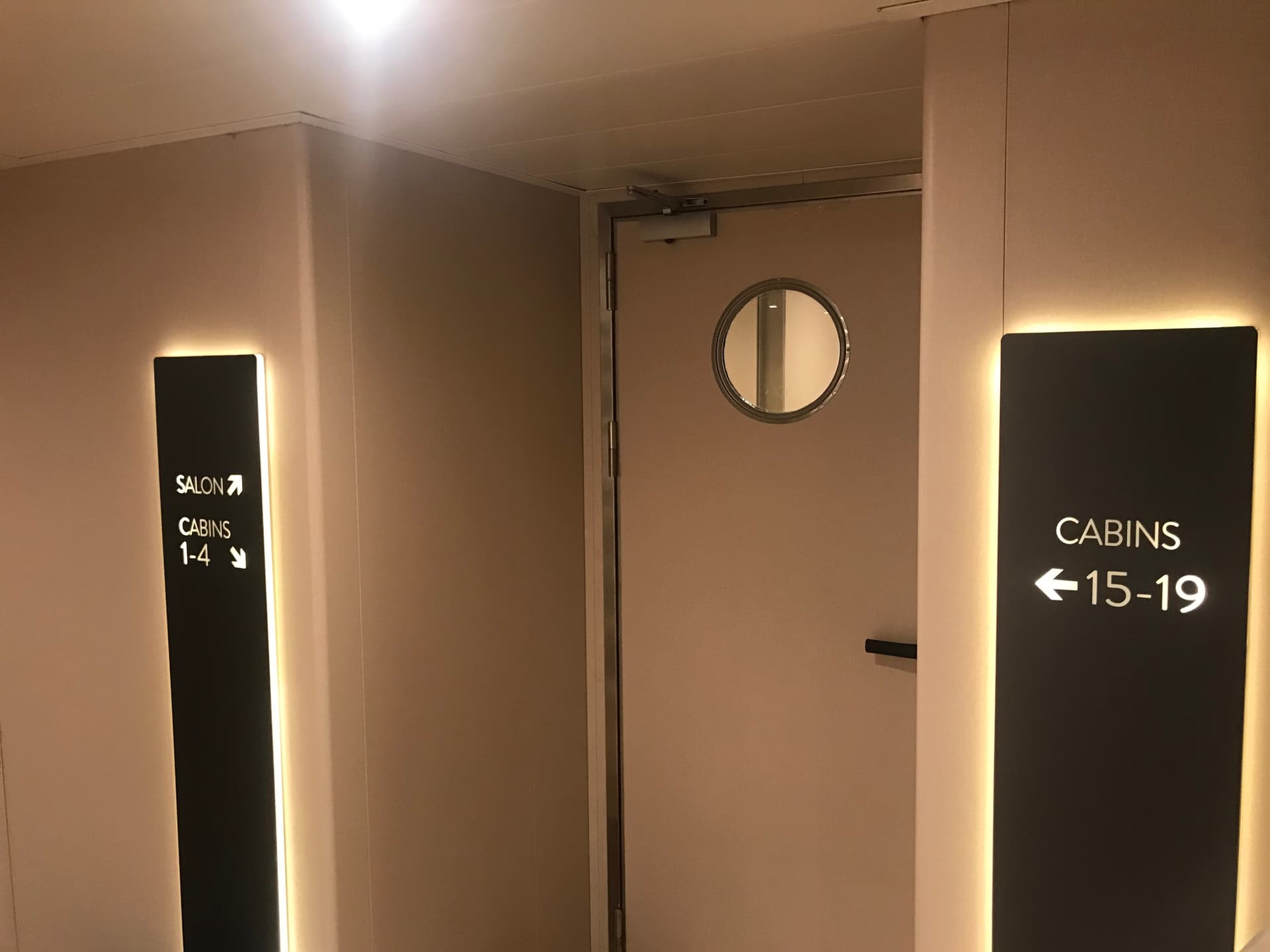 Ship hallway with illuminated directional signs for salon and cabins next to a door with a porthole window