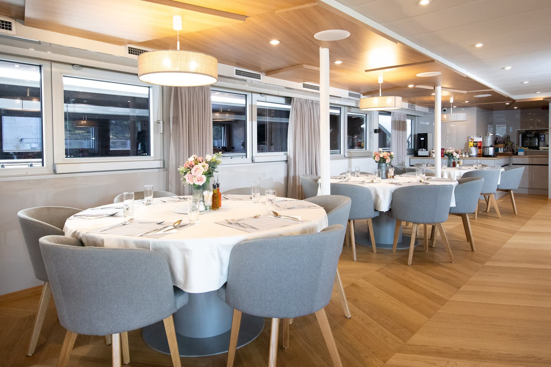 Modern restaurant interior with round dining tables, gray upholstered chairs, and wood ceiling paneling.