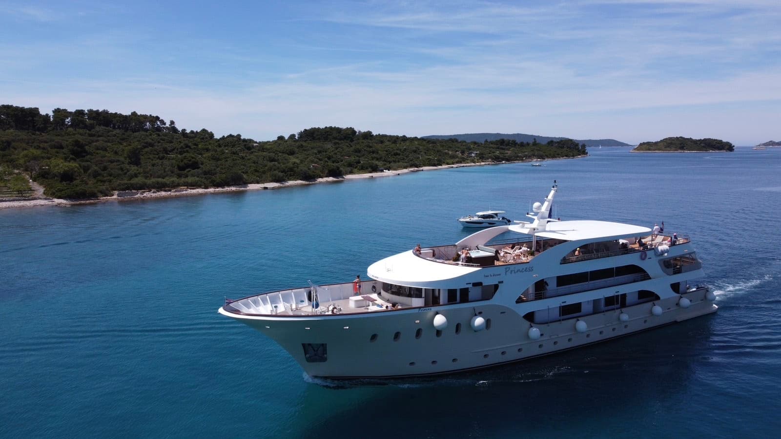Large white yacht cruising on blue coastal waters near forested islands