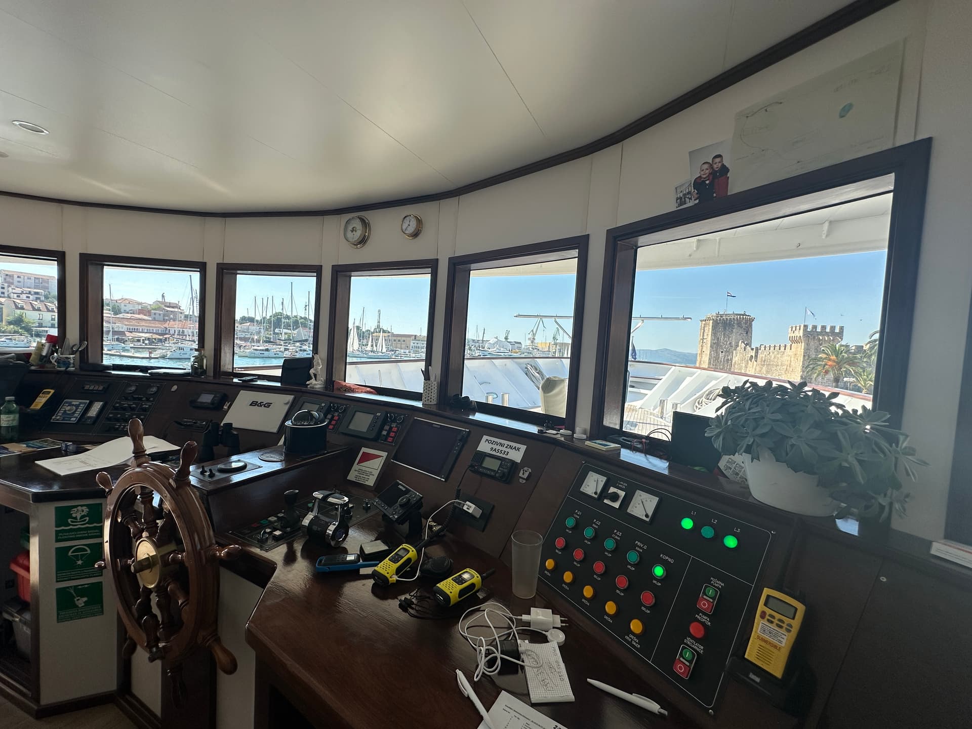 Ship's bridge console with wooden helm wheel and control panels overlooking a harbor and stone fortress