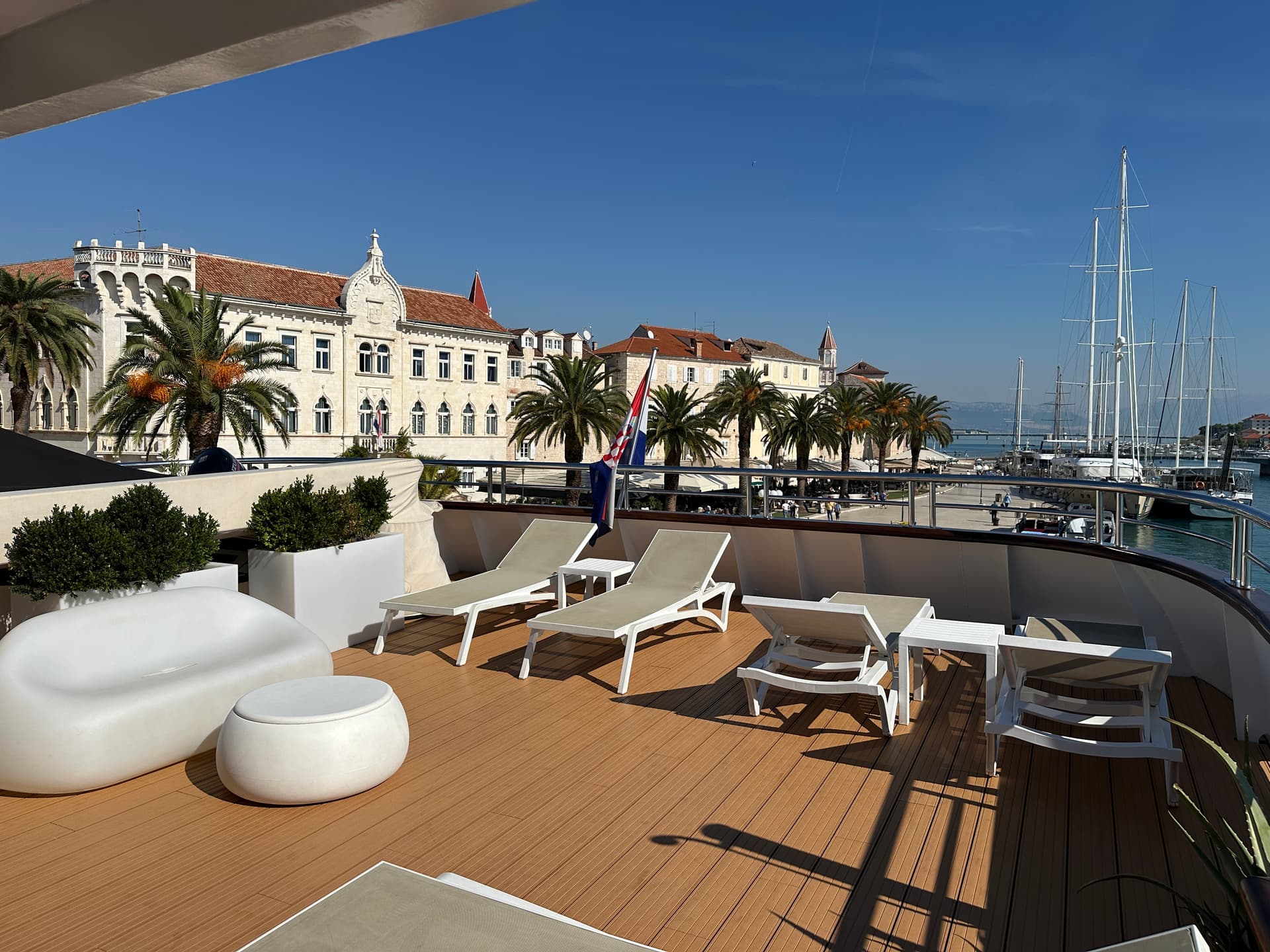Sun deck with loungers and white furniture overlooking a coastal harbor with historic buildings and palm trees
