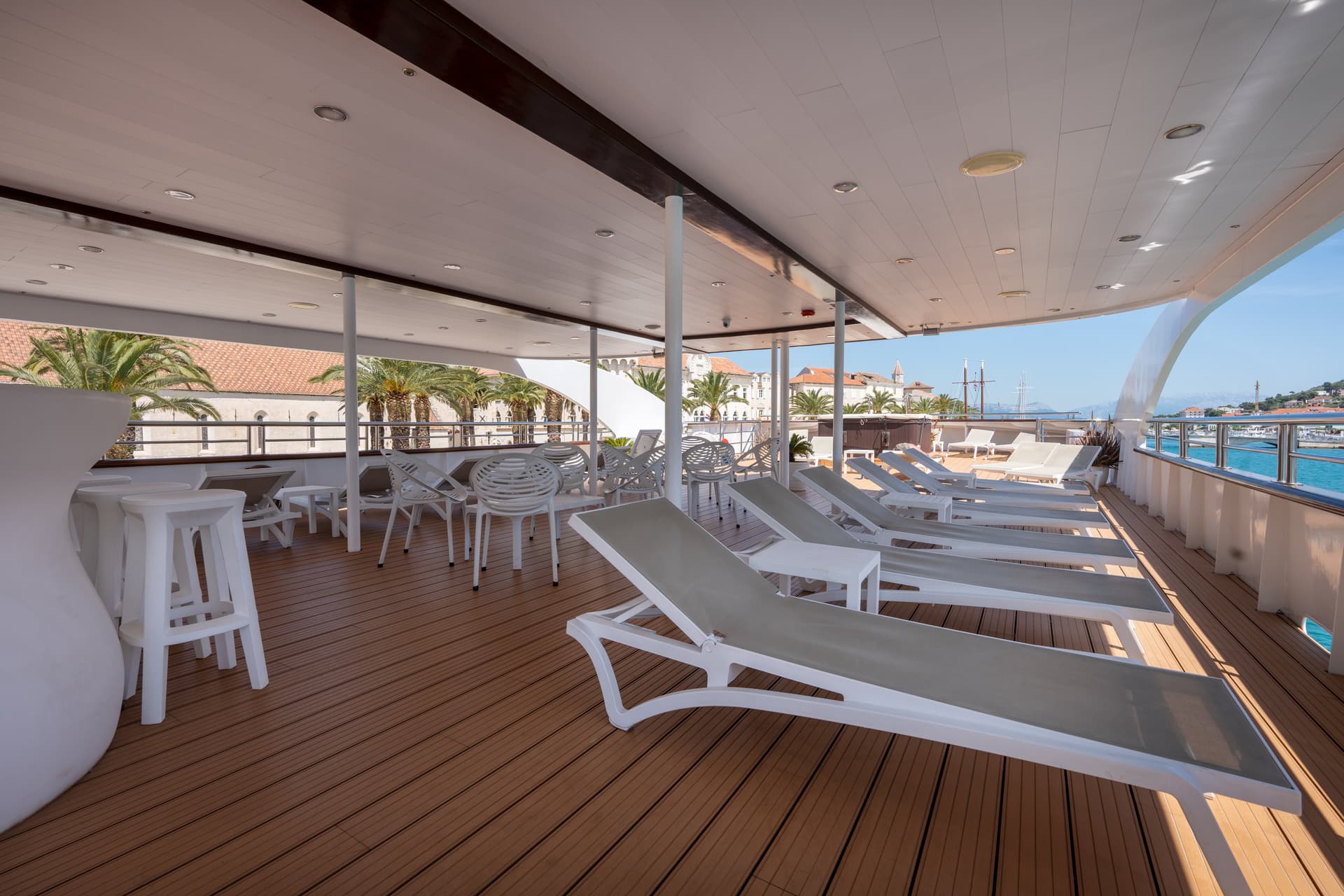 Sunny yacht deck with lounge chairs and bar seating overlooking coastal town harbor