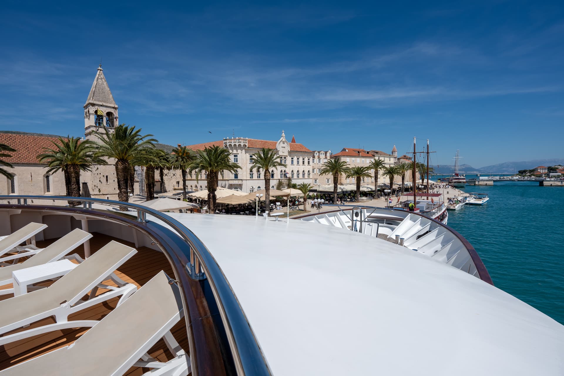 Lounge chairs on sunny cruise ship deck overlooking coastal town with palm trees and harbor