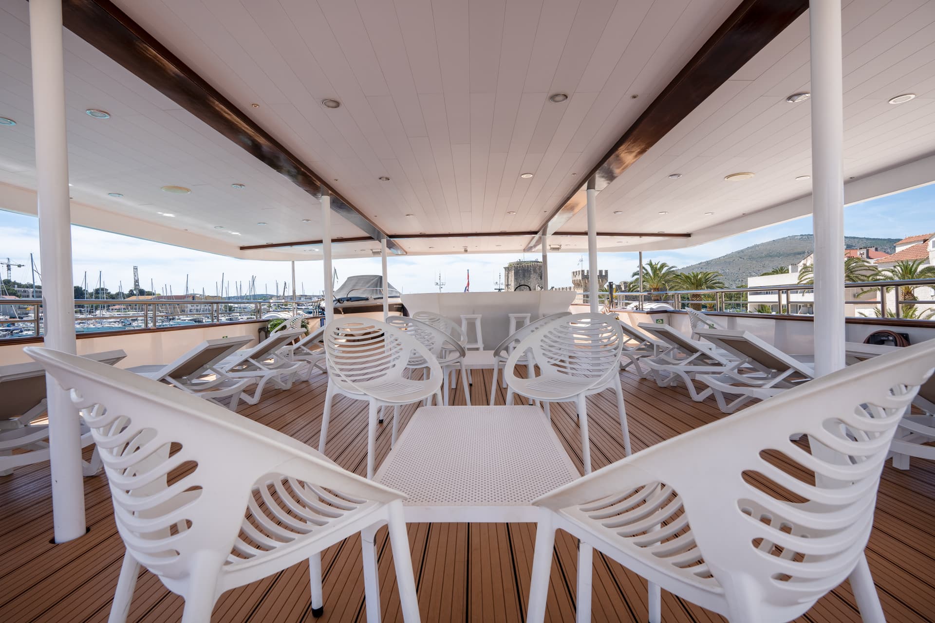 Covered deck with white chairs and lounge loungers on a boat with marina view