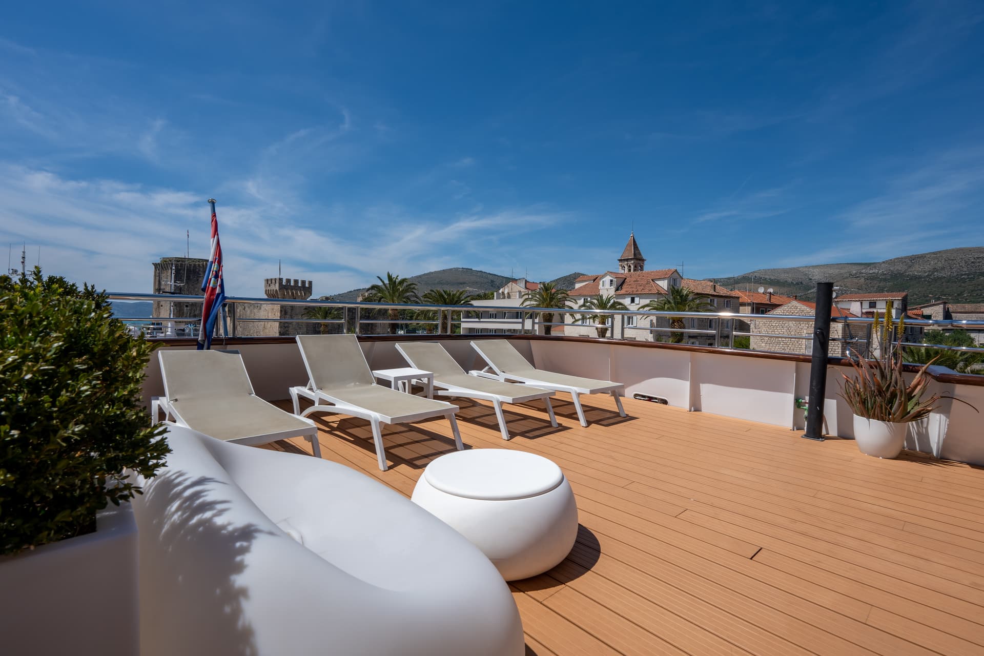 Sunny sun deck with lounge chairs and seating on a boat overlooking a coastal town with historic stone buildings and mount...
