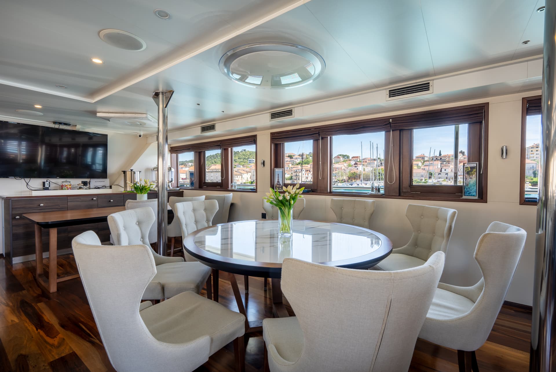Modern yacht dining area with round table and white chairs overlooking coastal town through windows
