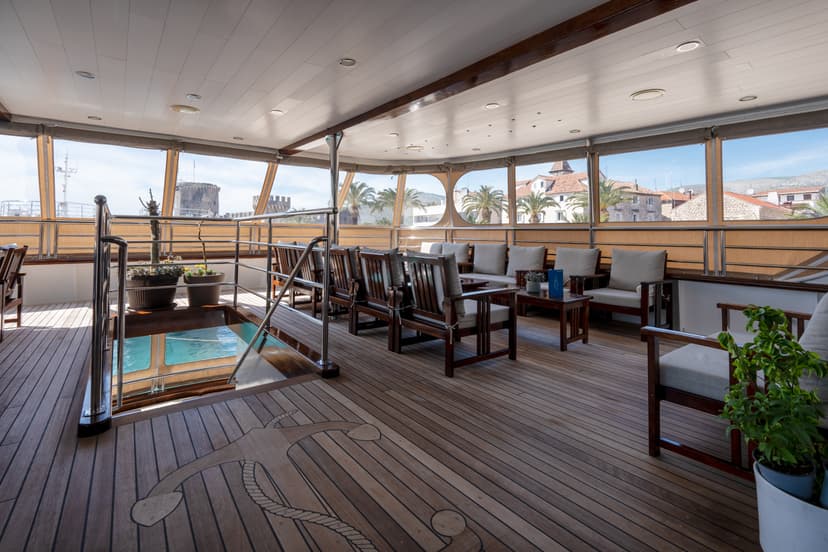 Lobby bar and restaurant seating area on a yacht deck with wooden flooring and large windows overlooking a coastal town.