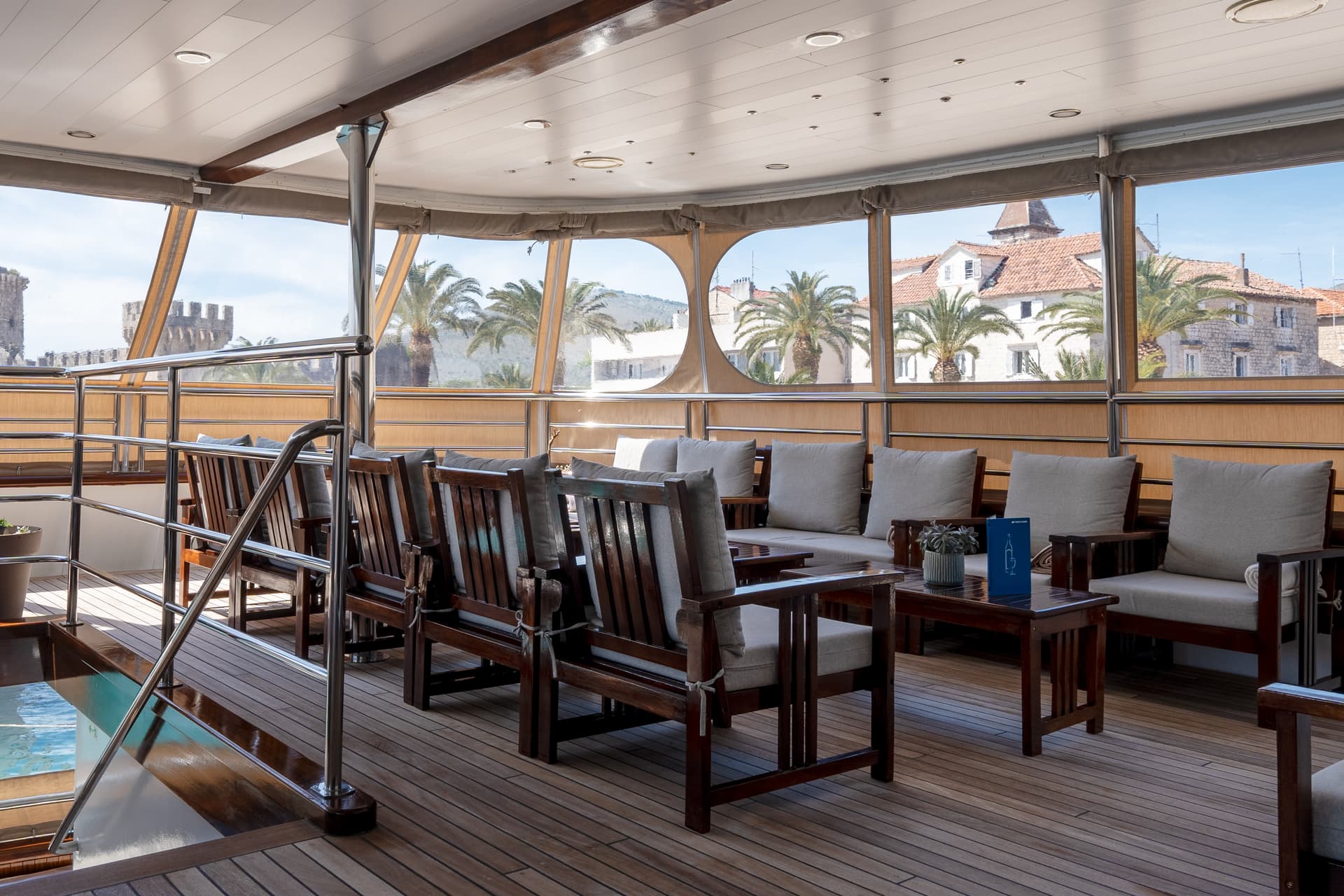 Lounge area on yacht deck with wooden armchairs and large windows overlooking coastal town