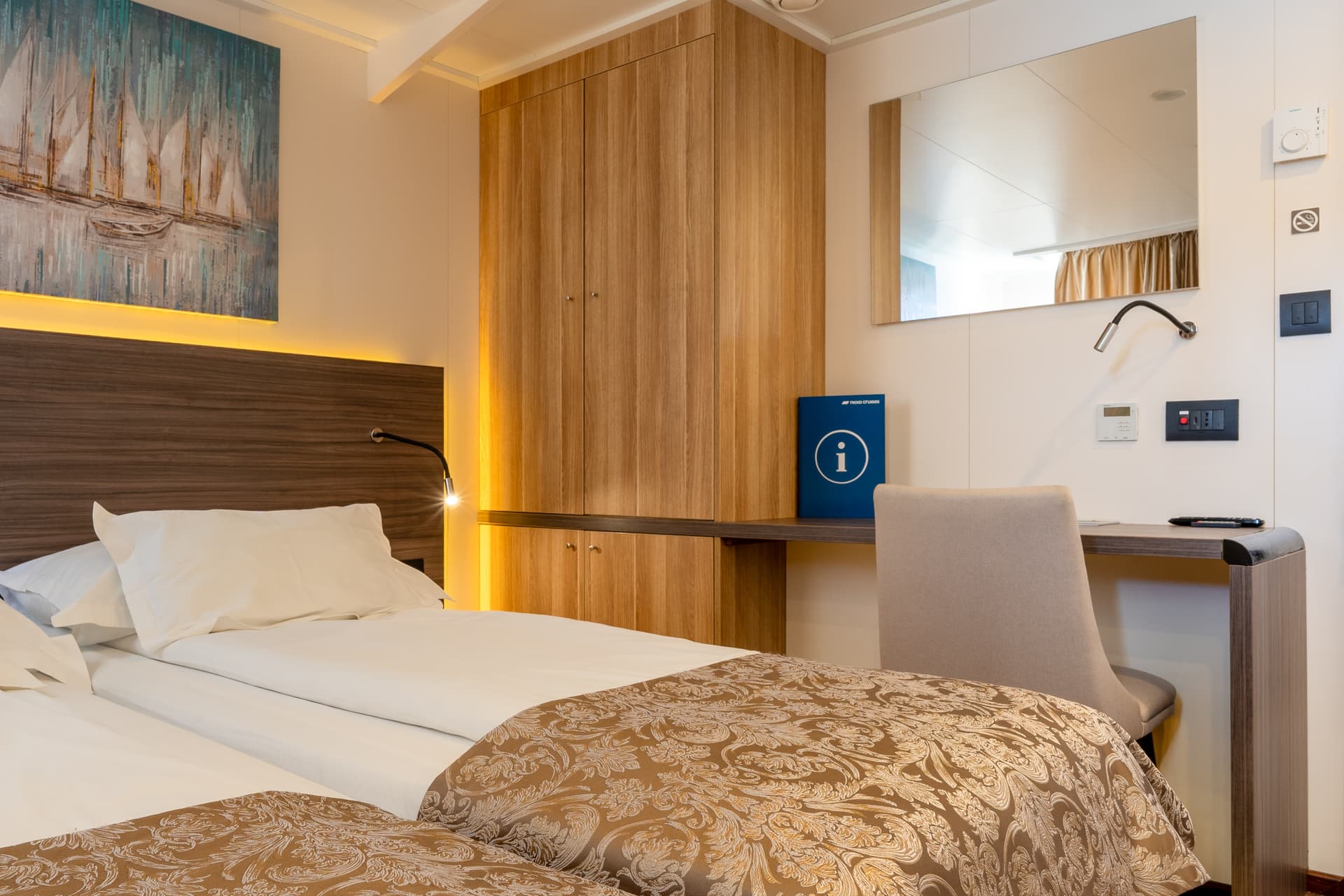 Cruise ship cabin with twin beds, wooden wardrobe, desk, and mirror