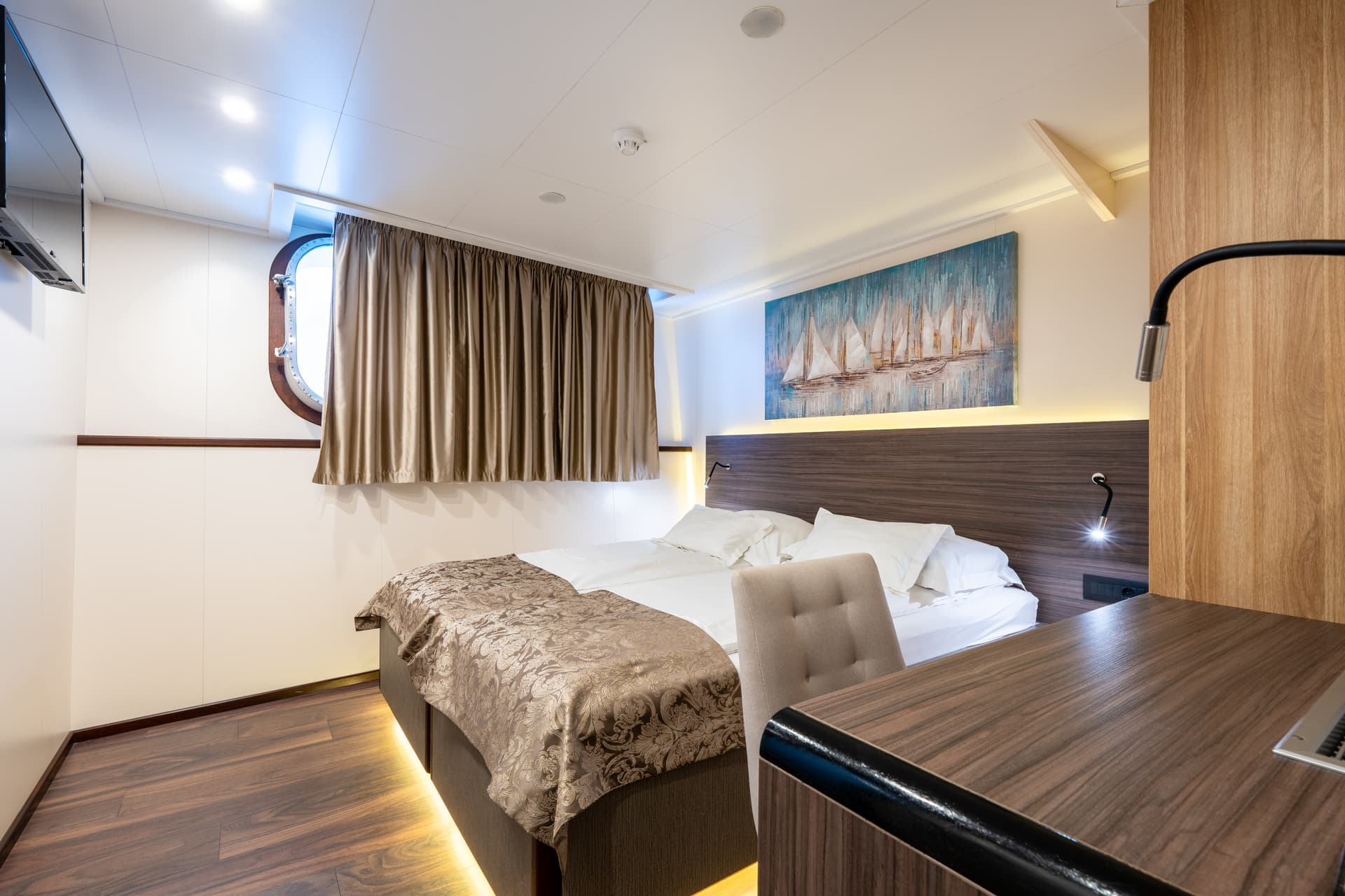 Ship cabin interior with double bed, porthole window, desk, and sailboat artwork