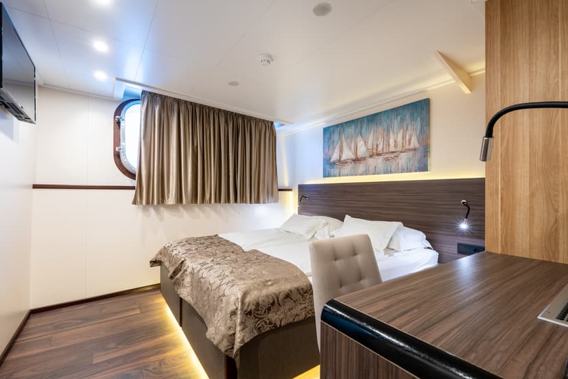 Ship cabin interior with double bed, porthole window, desk, and sailboat artwork