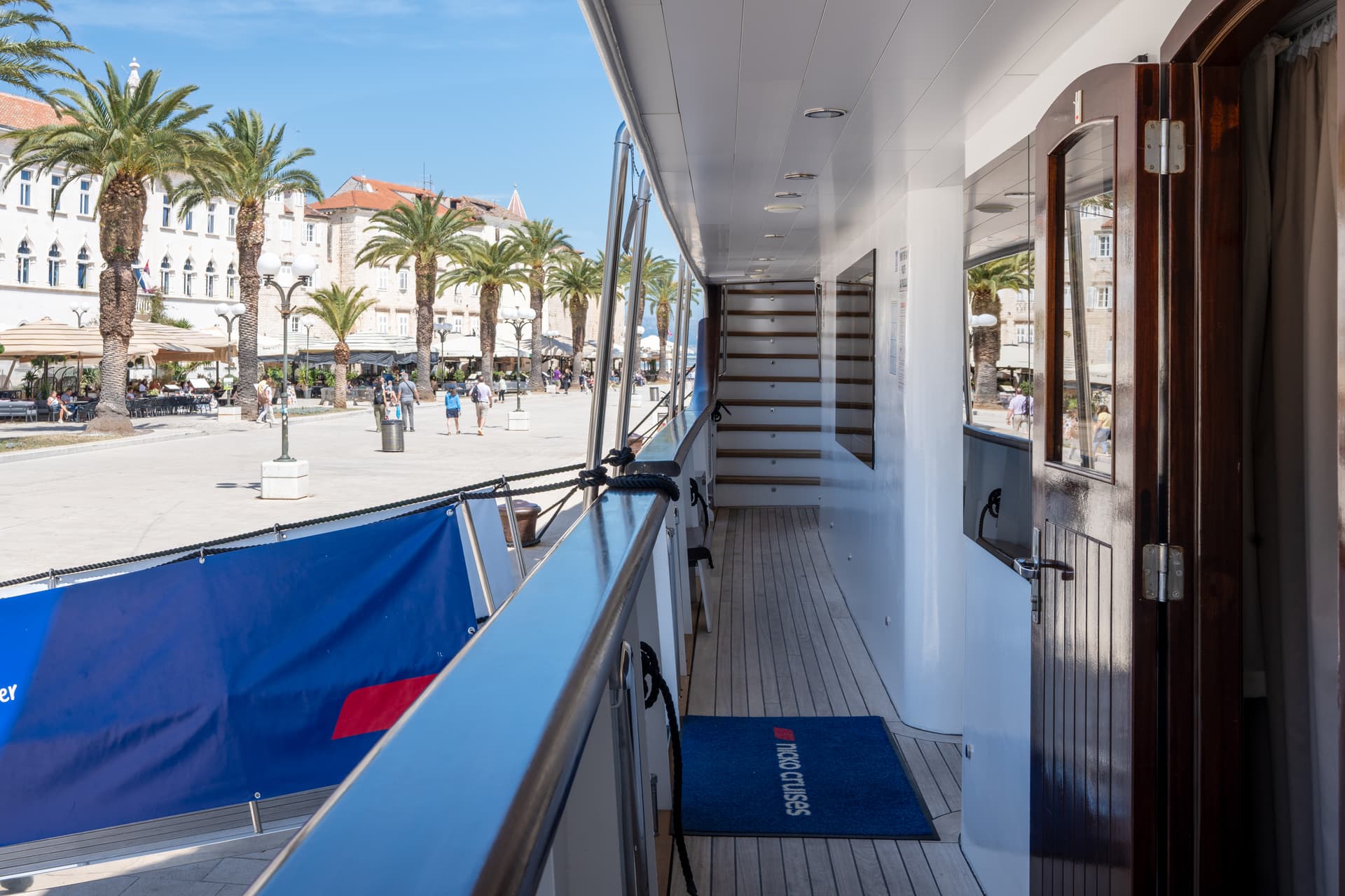 Cruise ship main deck exterior corridor with wooden flooring docked alongside a palm-lined promenade