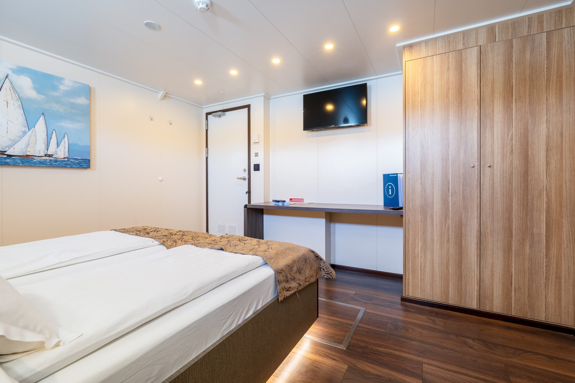 Standard deck cabin interior with bed, wooden wardrobe, desk, television, and sailboat artwork on wall
