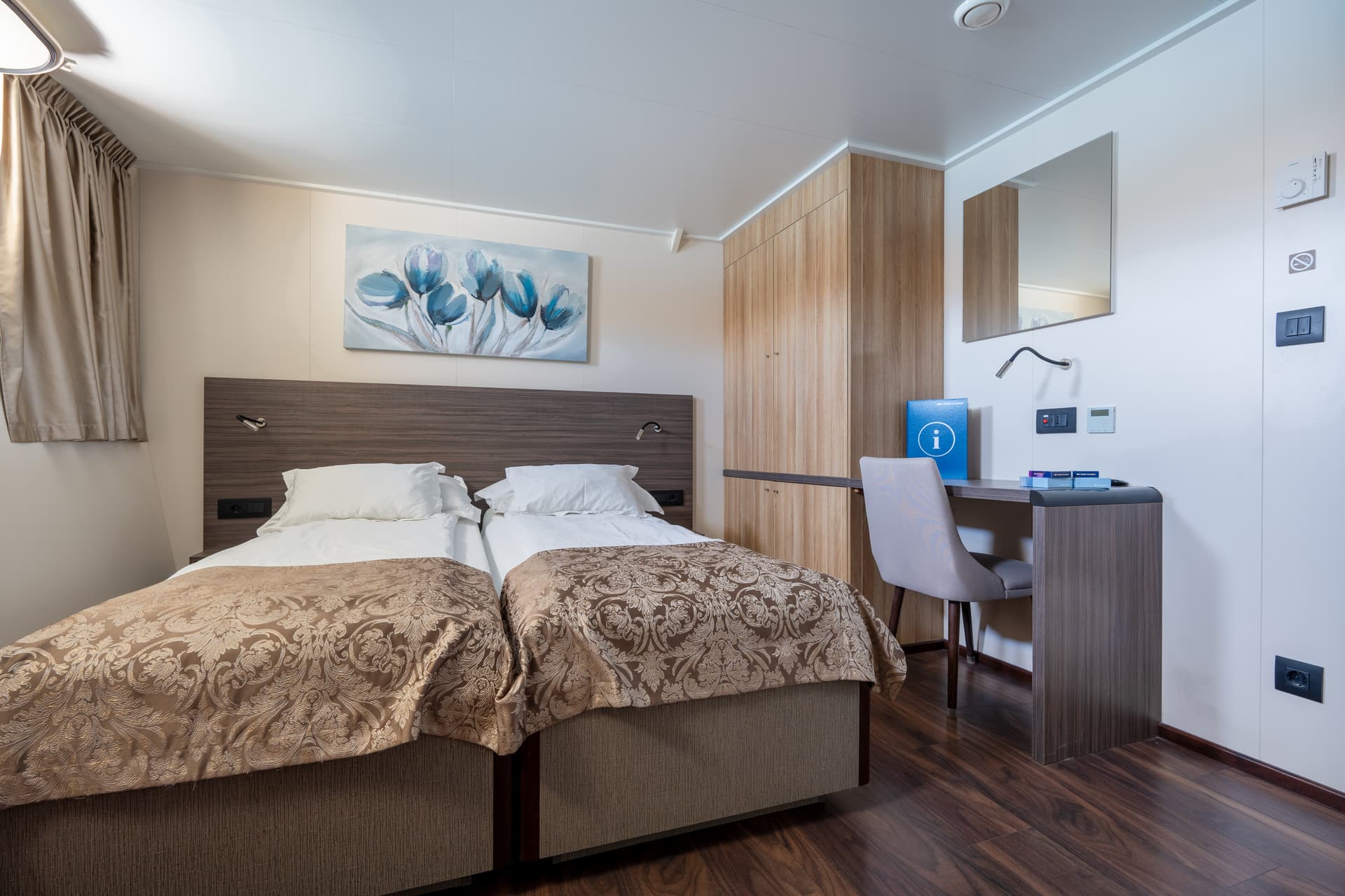 Cruise ship cabin with twin beds, wooden wardrobe, desk, and abstract floral wall art