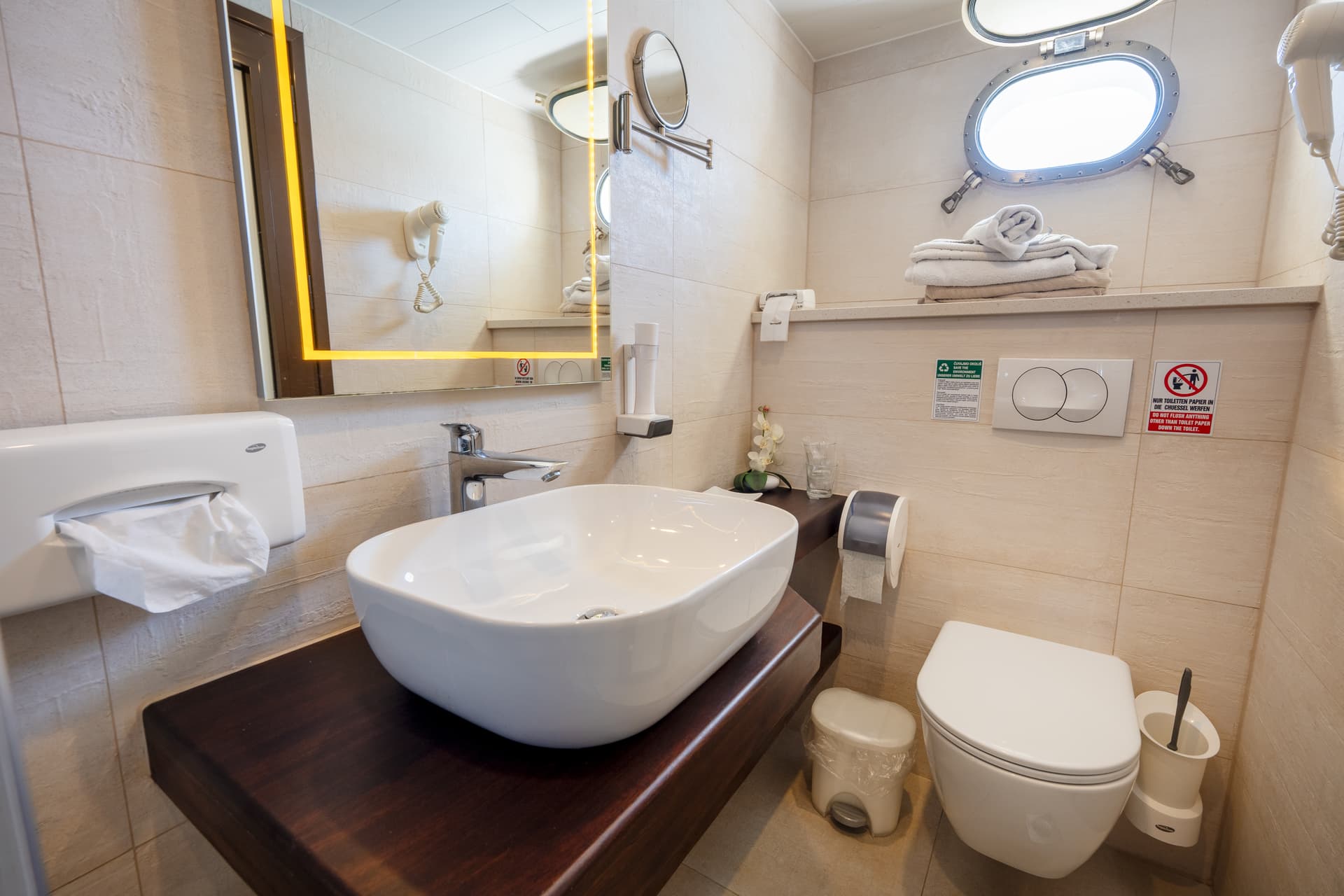 Modern compact bathroom with vessel sink, illuminated mirror, toilet, and porthole window