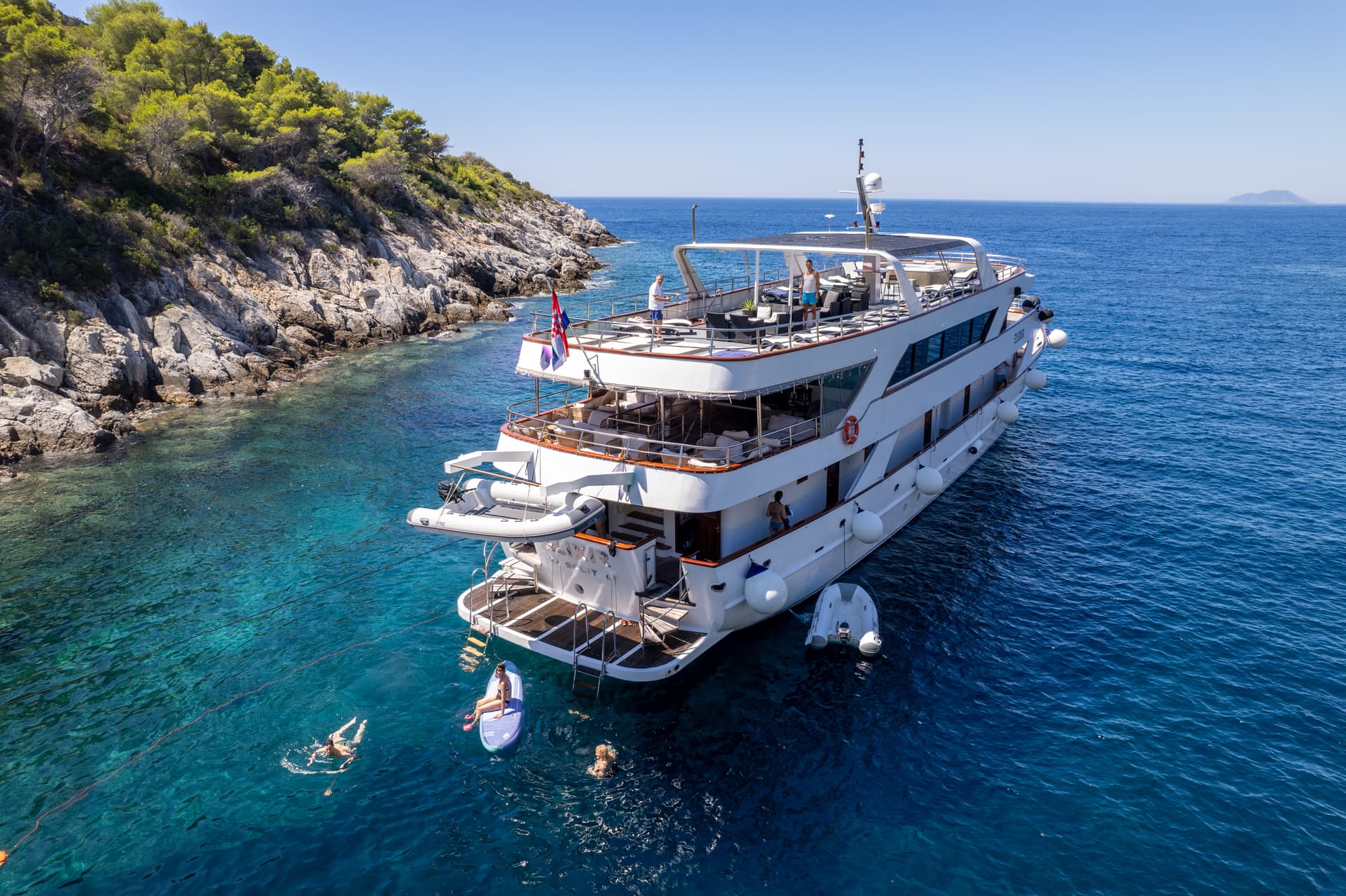 Luxury yacht anchored near a rocky coastline with people swimming in clear blue water