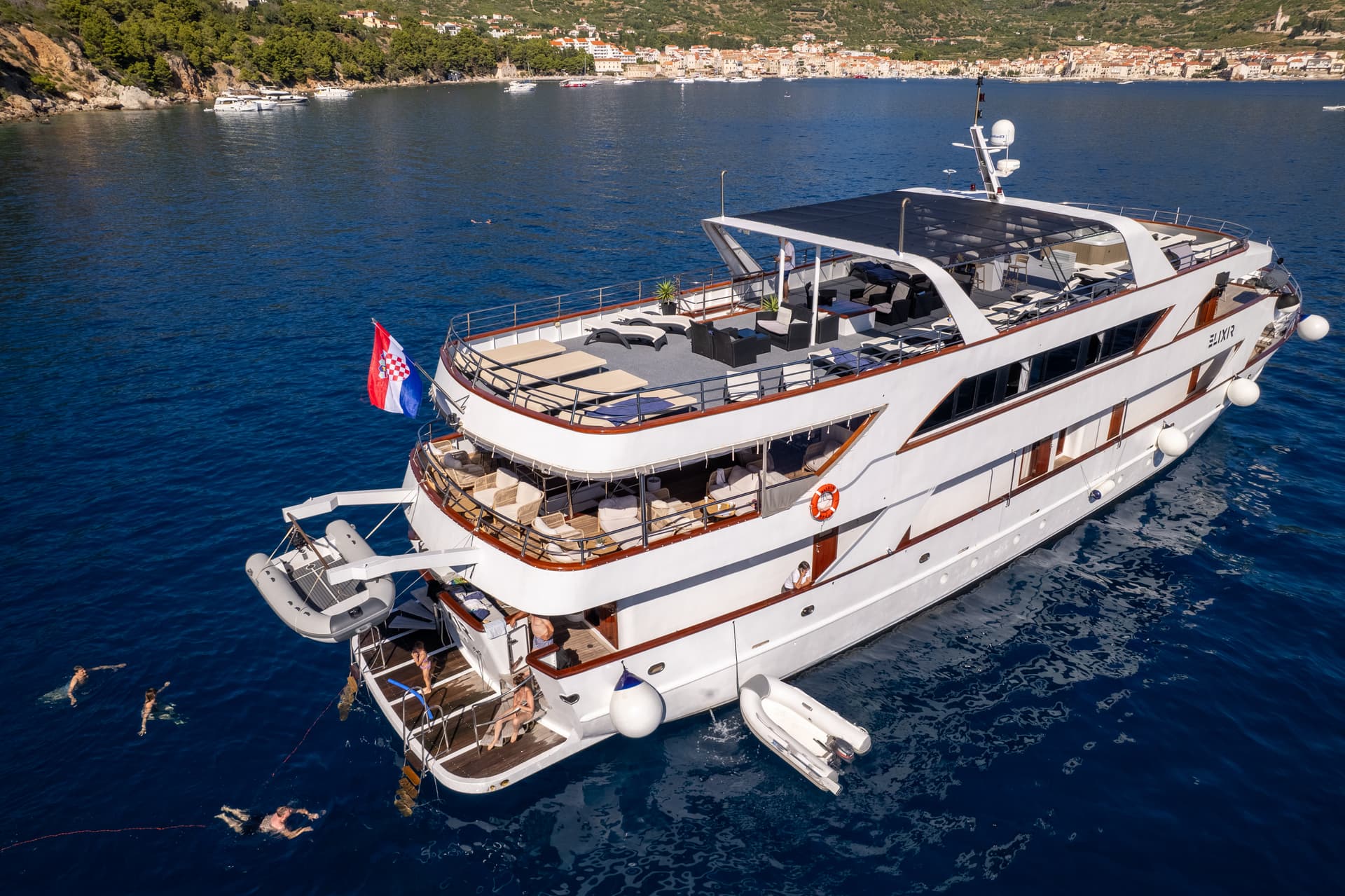 Large white yacht anchored in a blue bay with people swimming nearby and a coastal town in the background
